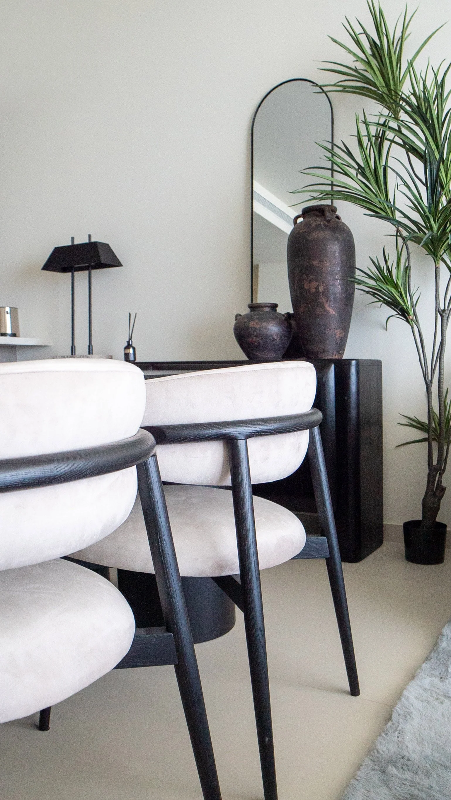 Modern living room with white chairs featuring black wooden legs, large dark vases on a black side table, a tall green potted plant, a black table lamp, and a mirror on the wall.
