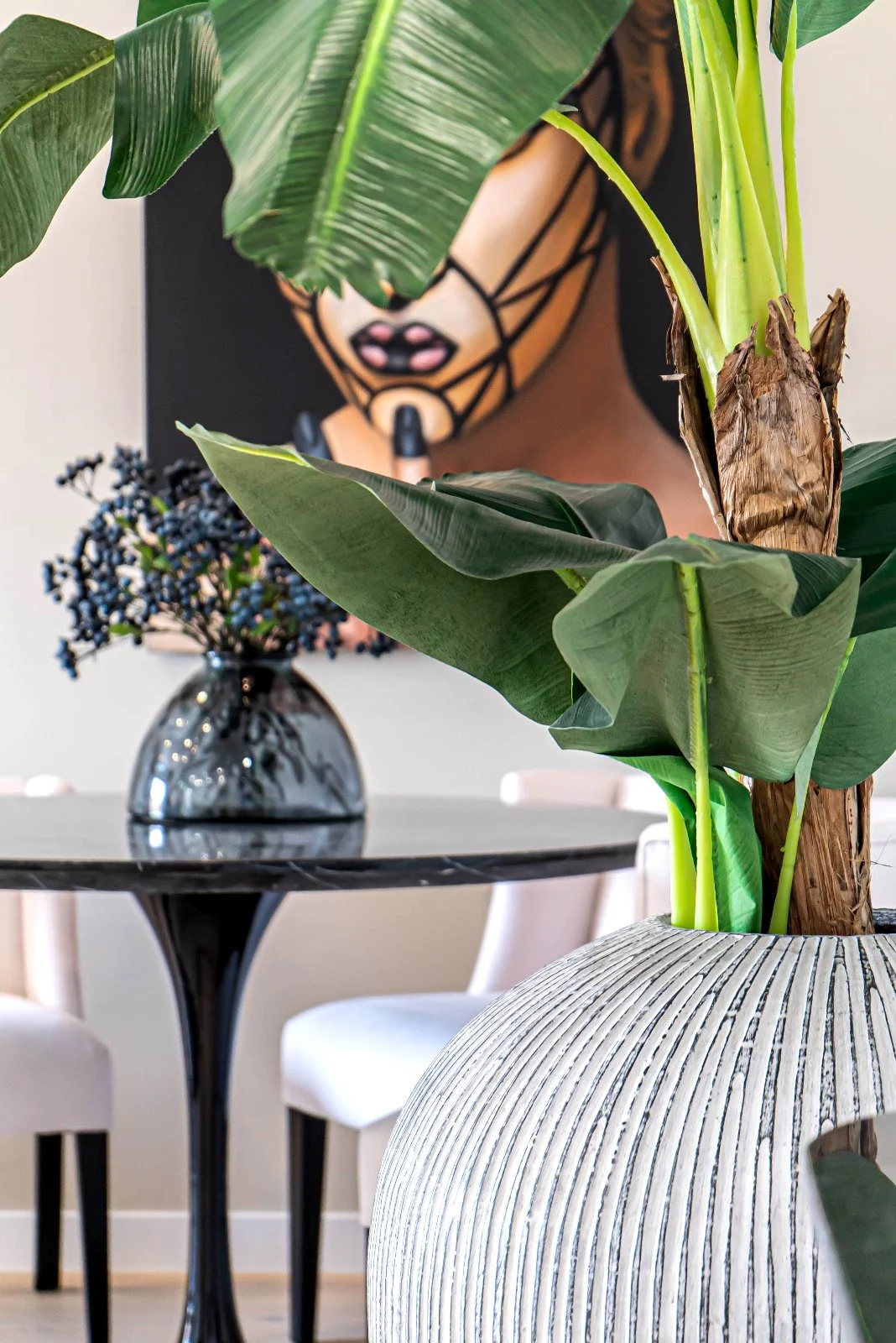 Interior space with large green leafy plant in a textured white and black striped vase placed on a table, with a black vase containing purple berries and a black and white geometric art piece in the background.