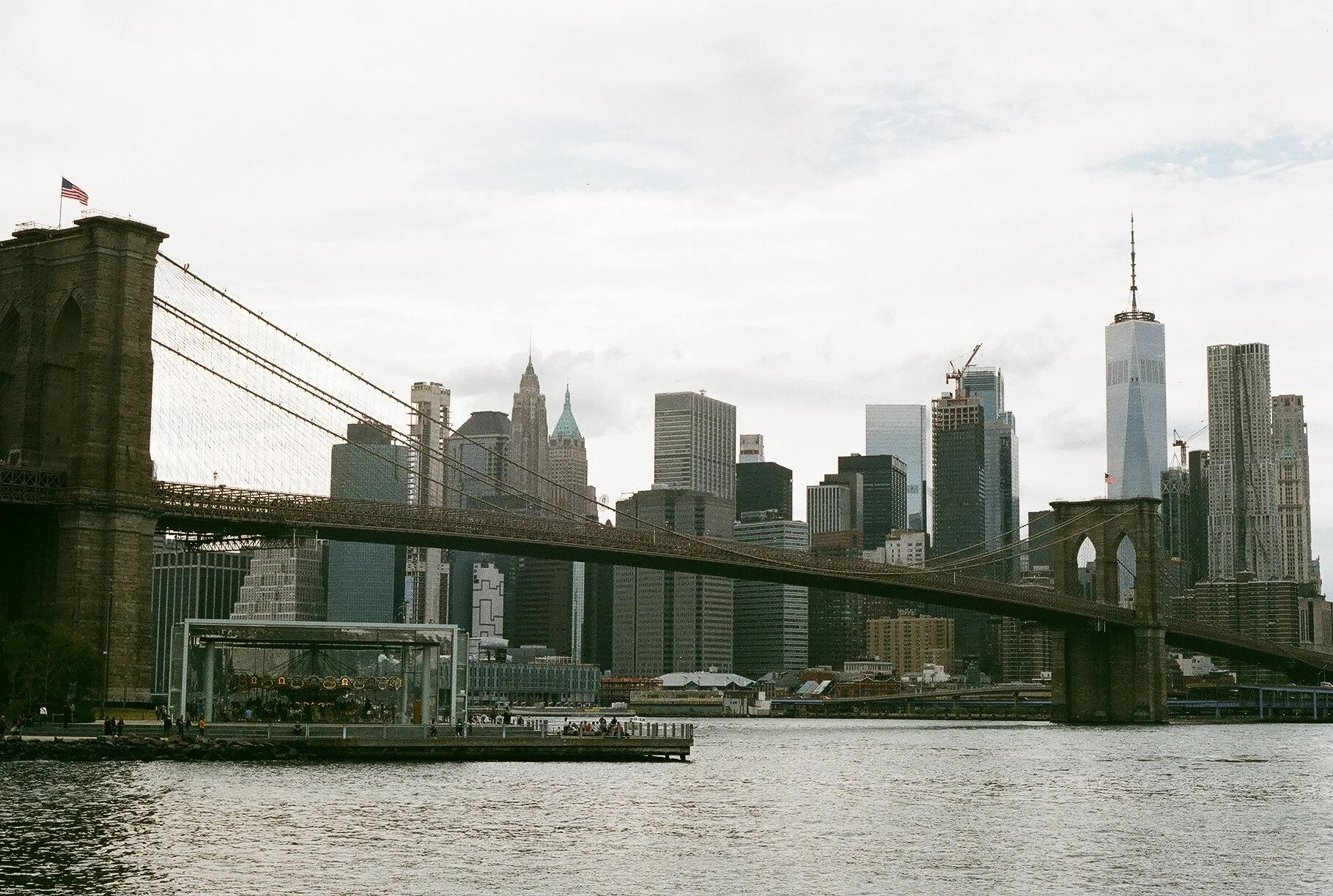 View of the New York City skyline with the Brooklyn Bridge in the foreground, spanning the East River, under an overcast sky.