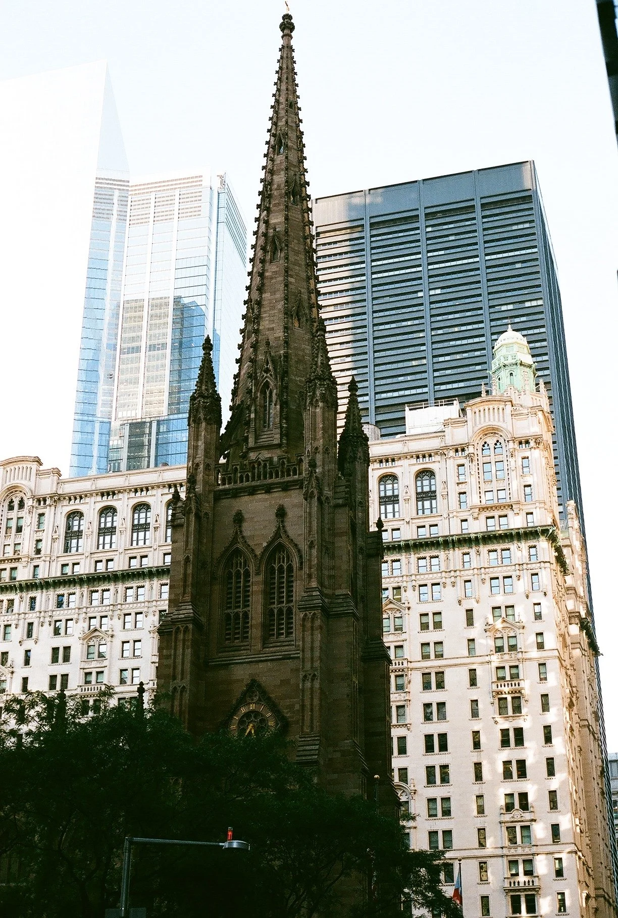A tall, dark Gothic-style church steeple surrounded by modern glass skyscrapers and historic buildings in an urban cityscape, with trees at the bottom.