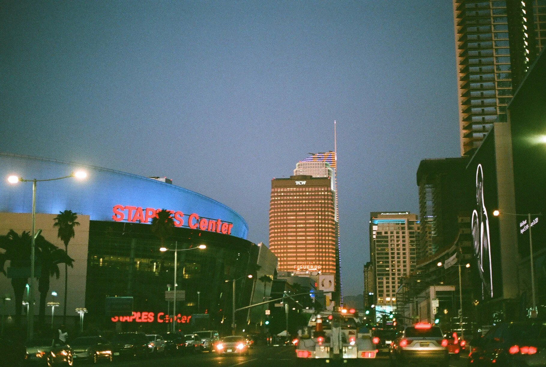 Nighttime cityscape featuring the Staples Center with its blue illuminated exterior and red signage, and tall buildings in downtown Los Angeles with lights.