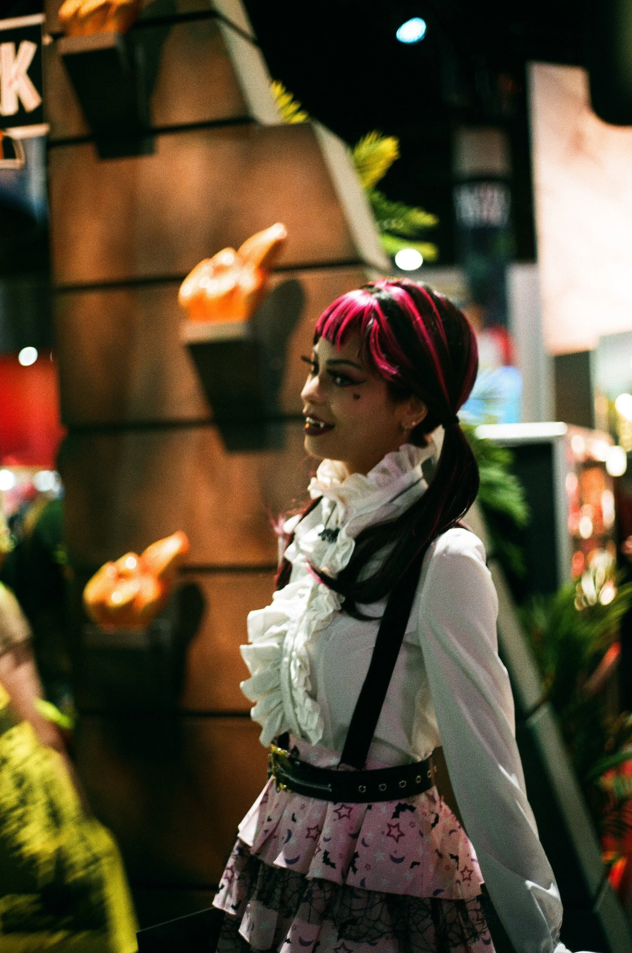 A woman with pink and black hair styled in pigtails, wearing gothic makeup, a white ruffled shirt, a pink skirt with black star and moon prints, and black suspenders, standing in a dimly lit setting with illuminated decorations in the background.