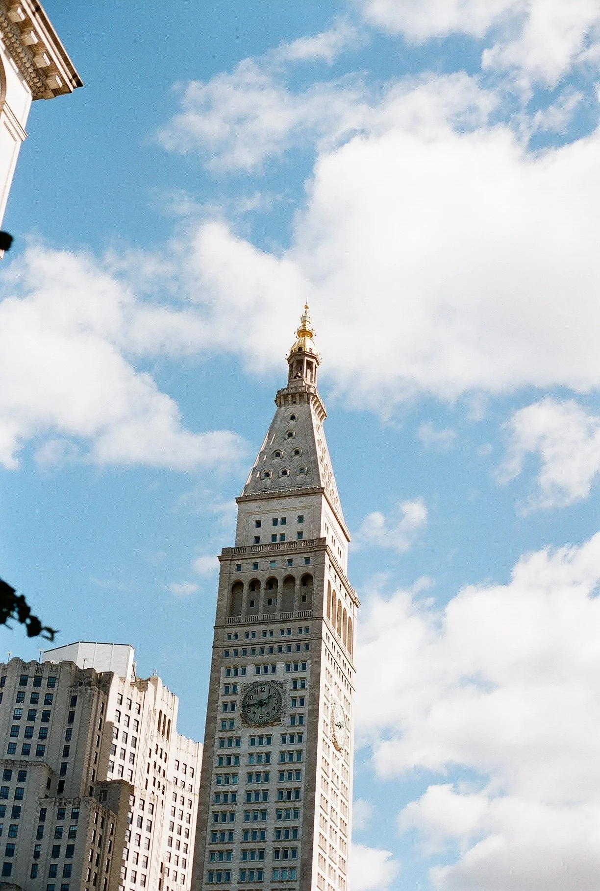 Tall clock tower with a pointed, ornate top, set against a blue sky with scattered clouds, in an urban area.