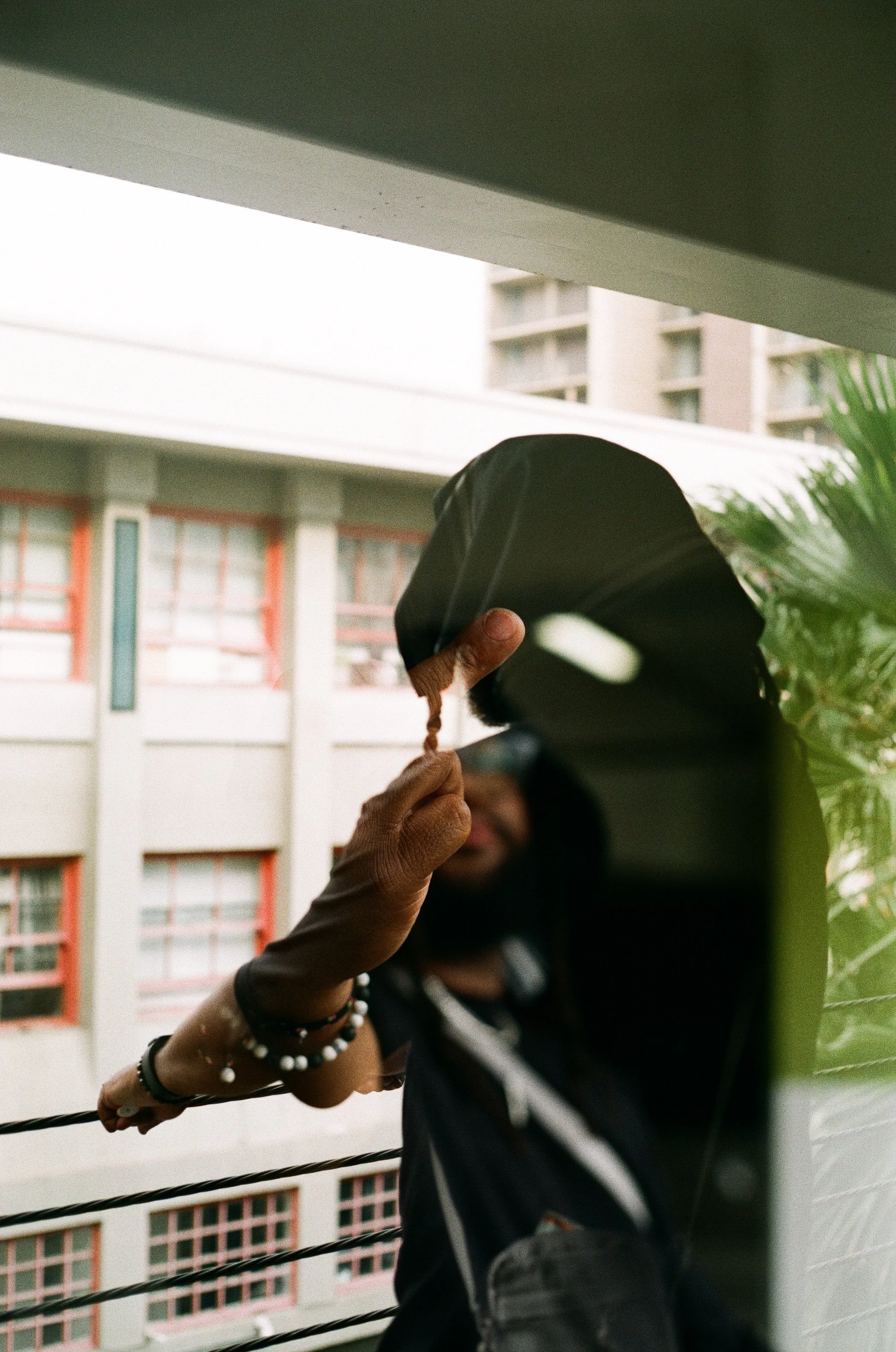 Person wearing a hoodie and bracelets, with a reflection visible in a mirror, standing outdoors near a balcony with buildings in the background.