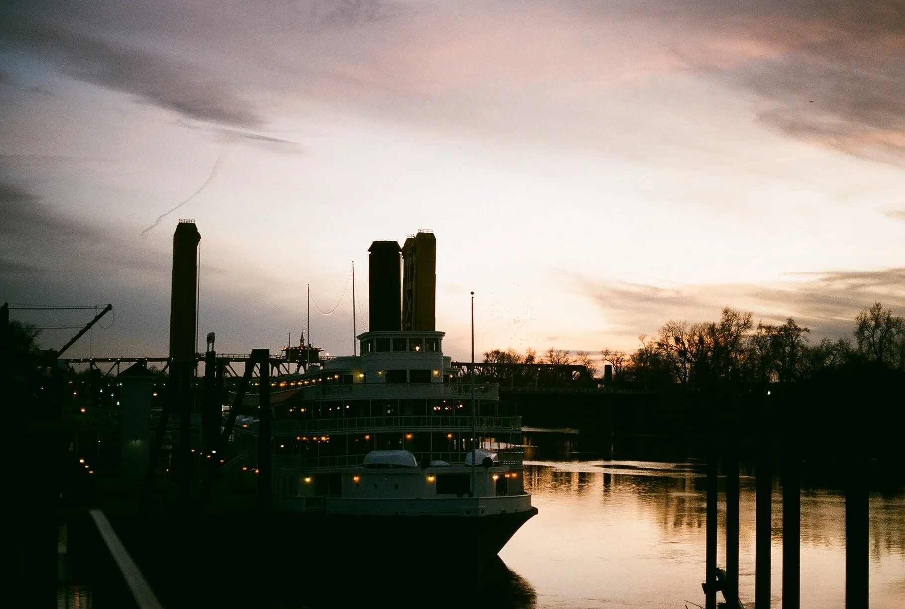 Silhouette of a riverboat docked on the water at sunset, with trees and a bridge in the background.