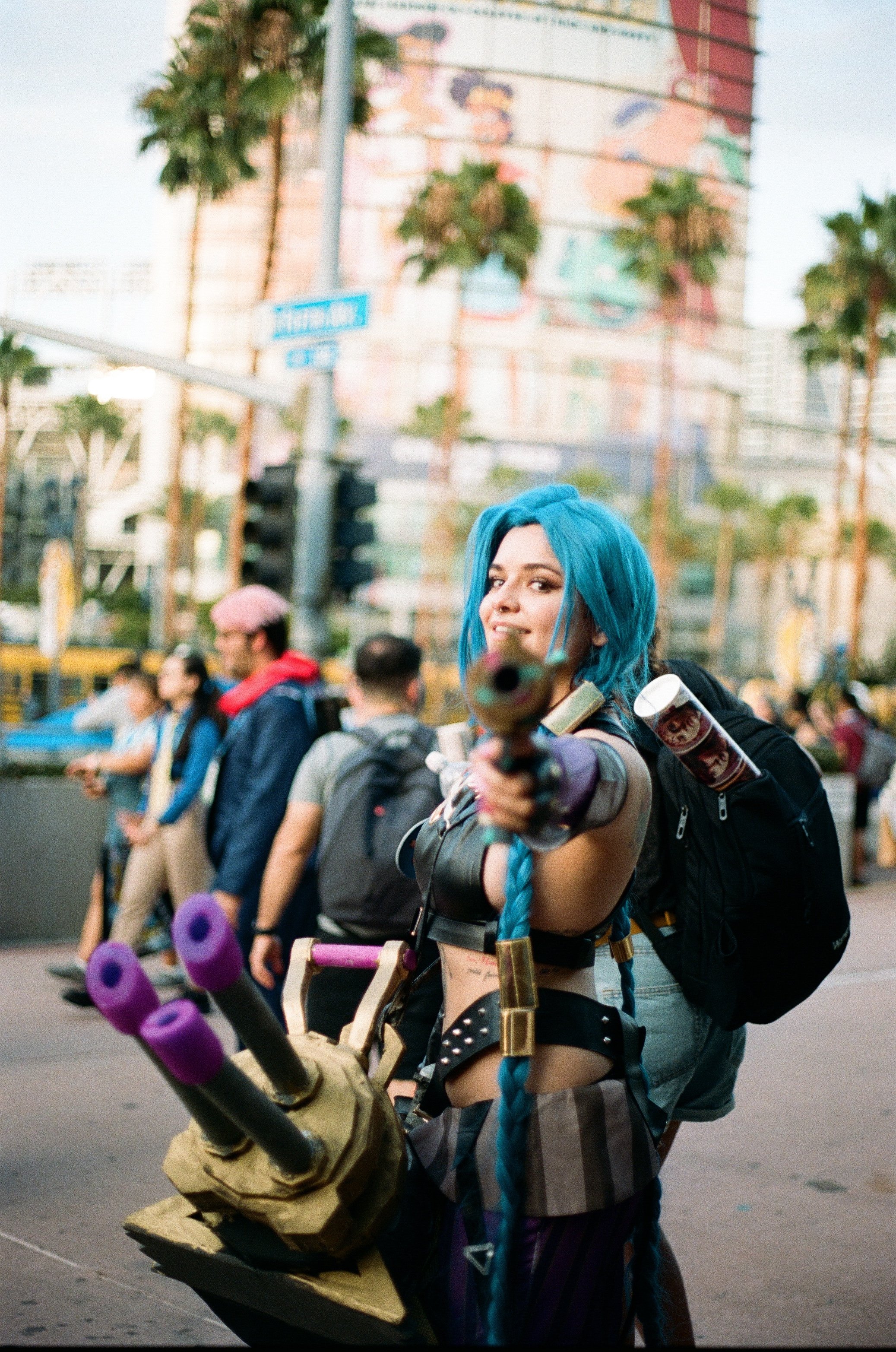 Woman with long blue braided hair pointing a toy gun at the camera, dressed in cosplay costume with a backpack and prop weapons, standing on a city street with pedestrians and tall buildings in the background.