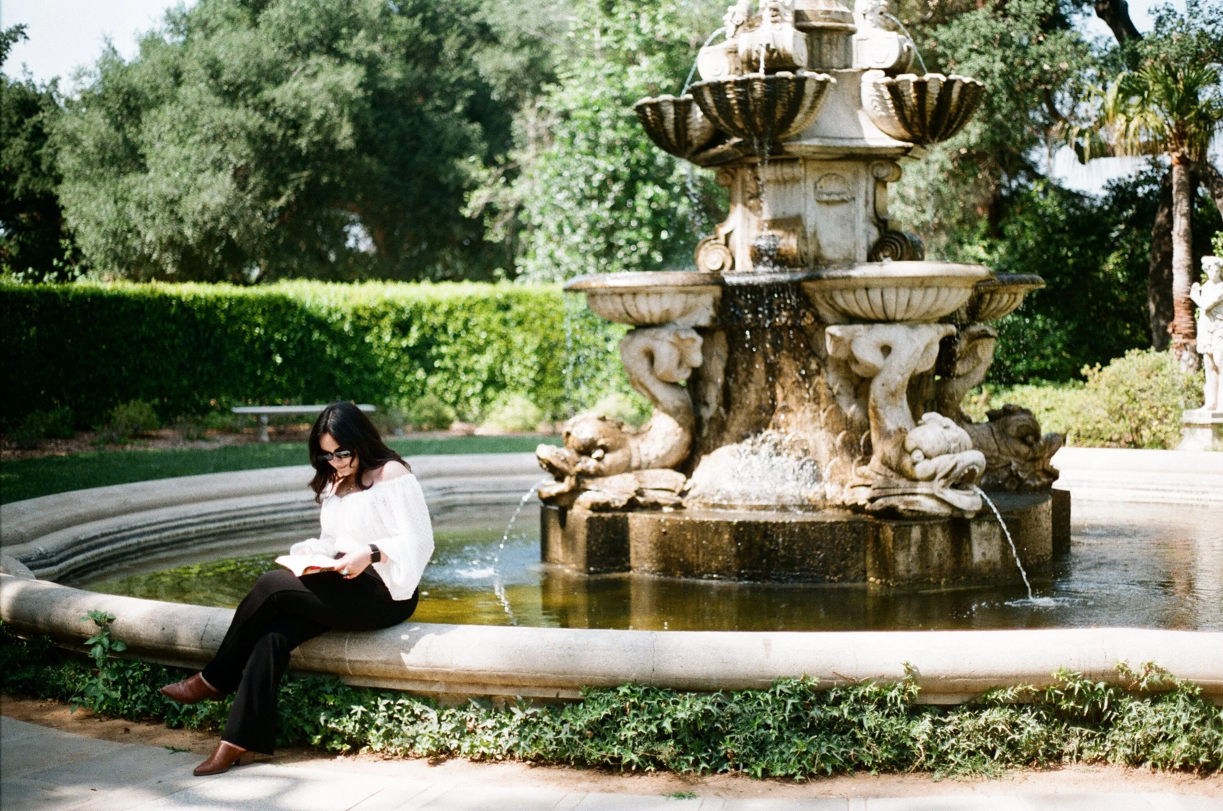 A woman sitting on the edge of a fountain in a park, reading a book on a sunny day, surrounded by lush greenery.