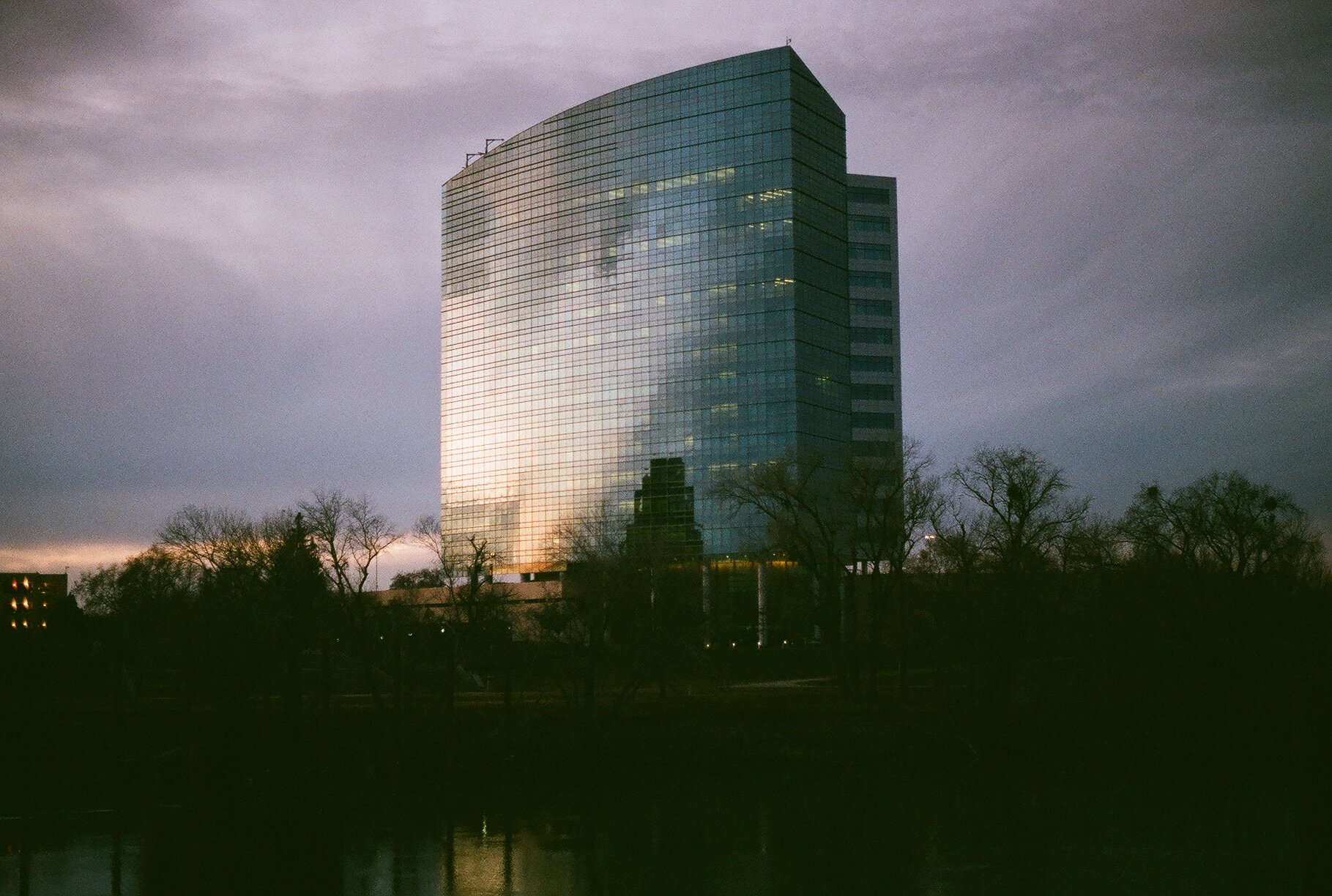 A tall modern glass office building reflecting the cloudy sky and nearby trees, with some windows lit up, surrounded by leafless trees in the foreground during dusk or dawn.