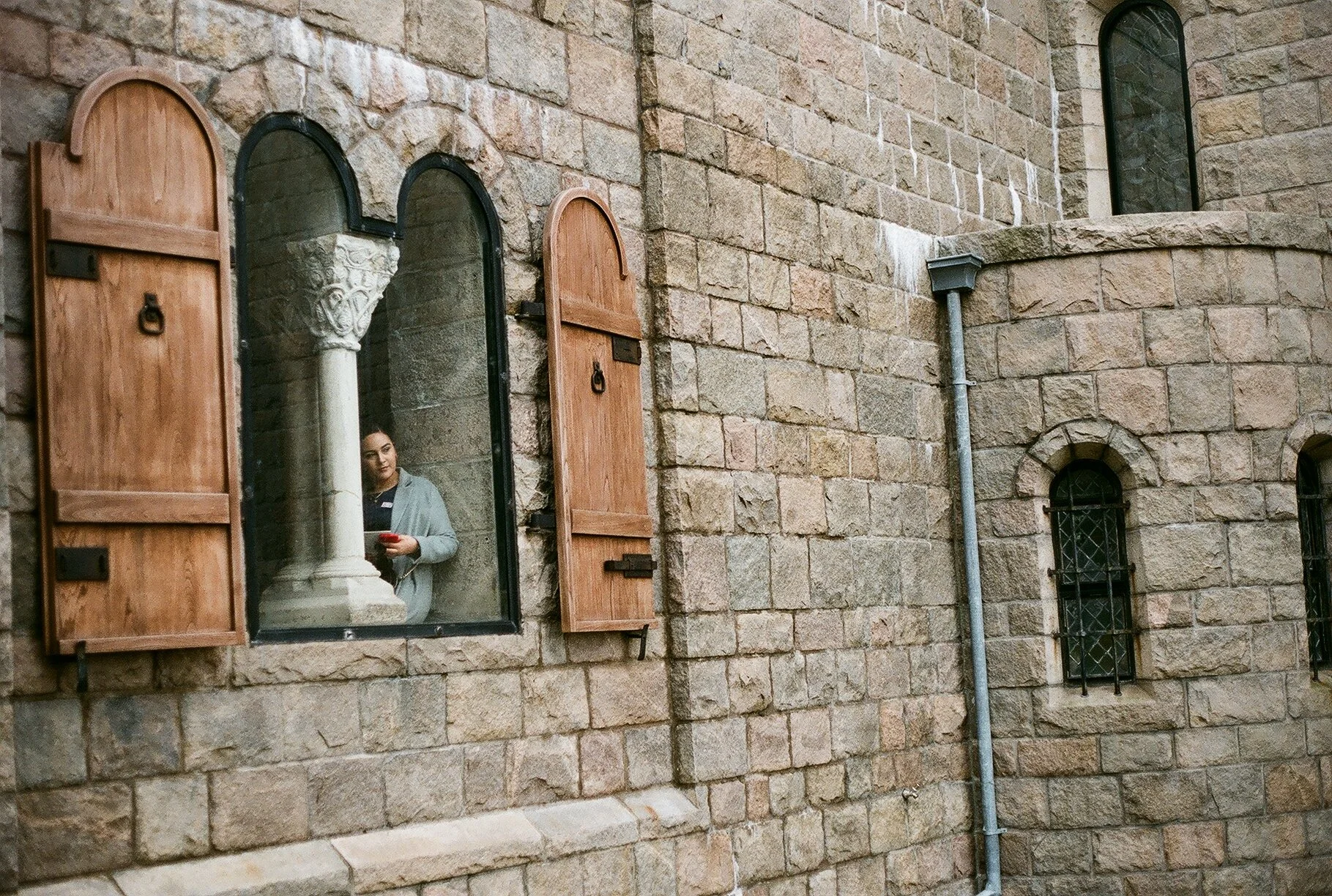 A woman looking at her phone through a window with open wooden shutters on a stone building. The window features a decorative stone column inside.