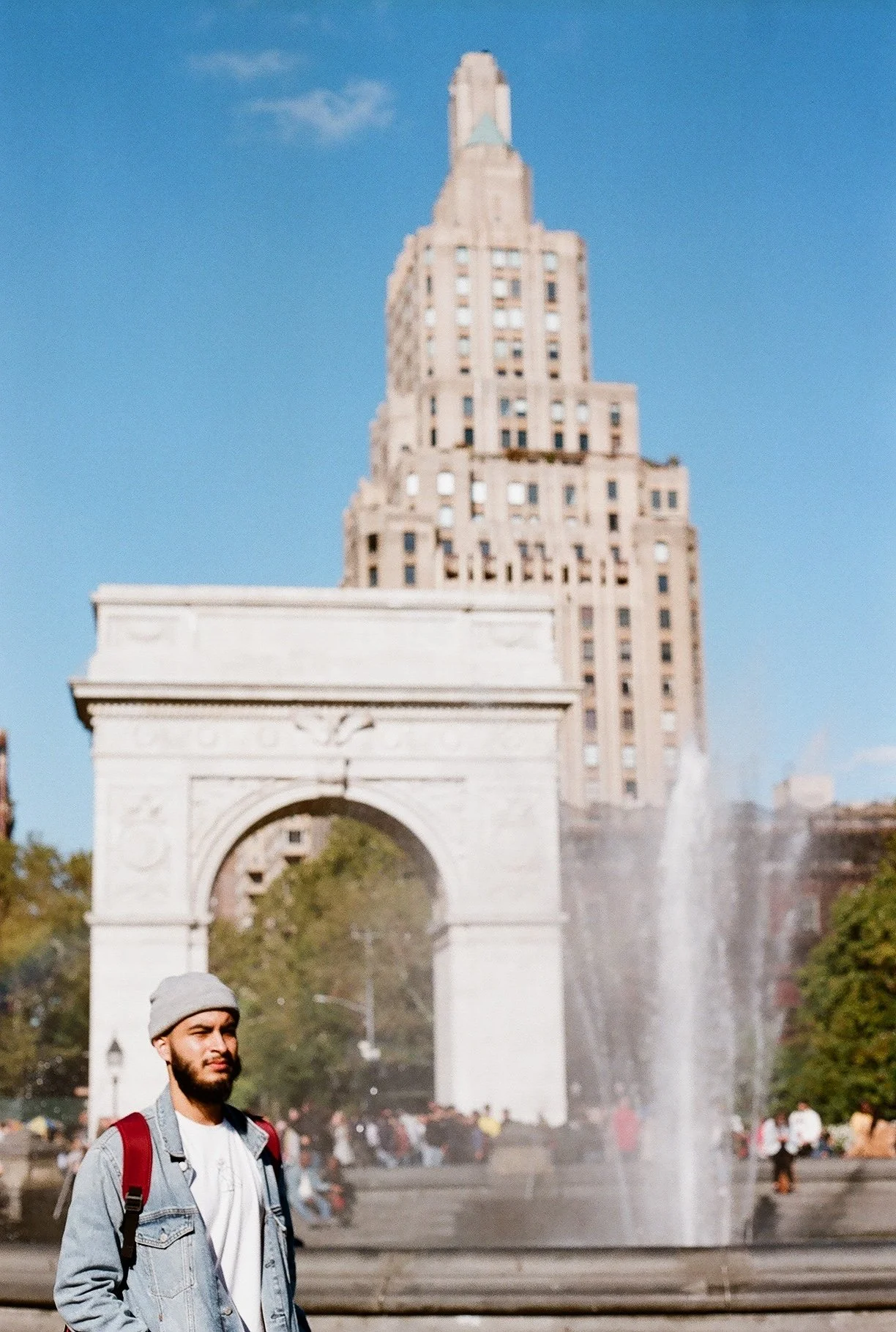 A man with a gray beanie, denim jacket, and backpack standing in front of a fountain with water spraying, with the Washington Square Arch and a tall building in the background.