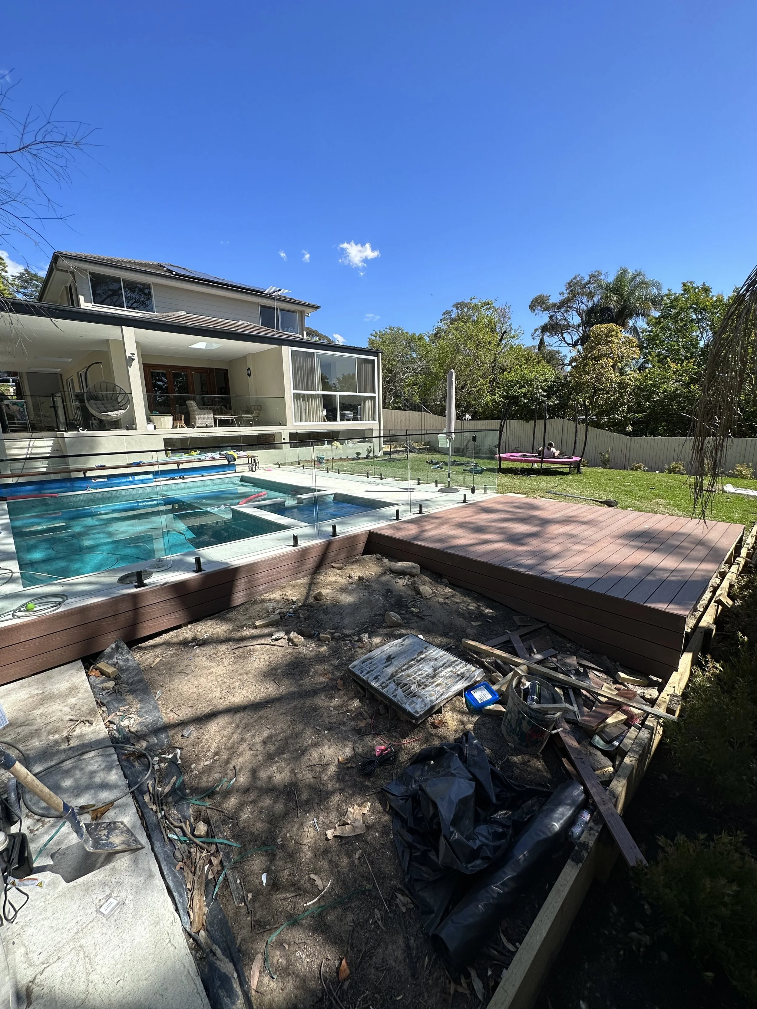 Backyard with a house, an in-ground pool under construction with a glass fence, a completed wooden deck, and a grassy area with trees and a pink trampoline, on a bright sunny day.