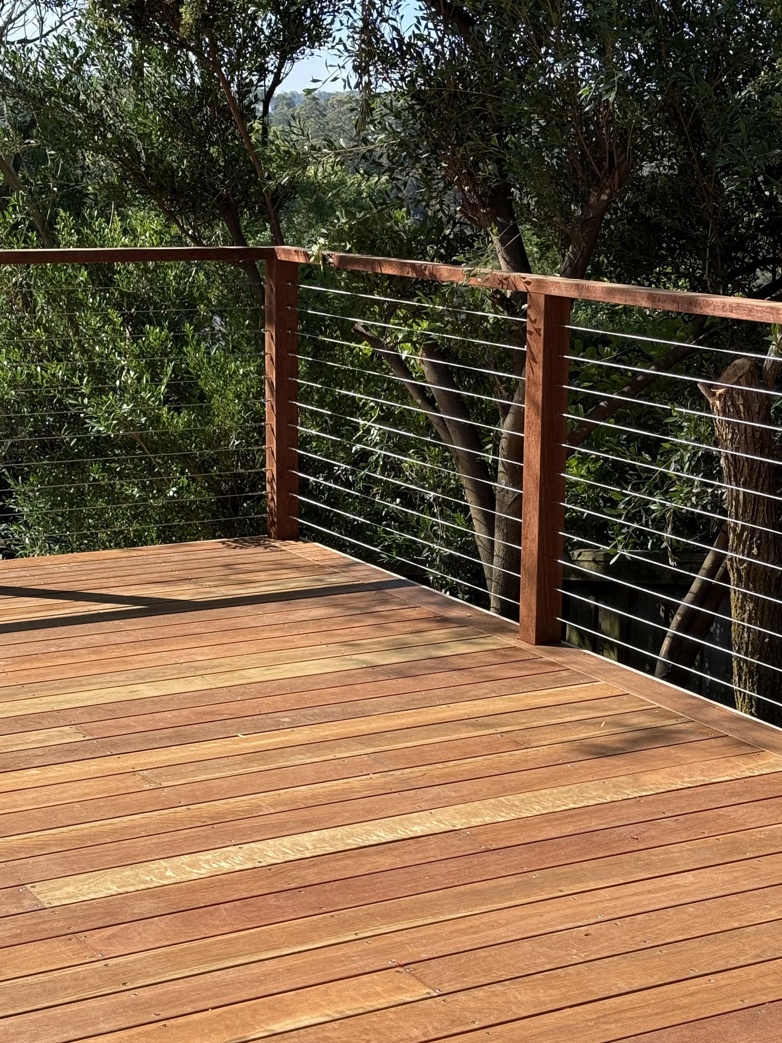 A wooden deck with a railing made of vertical wood posts and horizontal metal wires, overlooking a green leafy tree and a hill in the background.