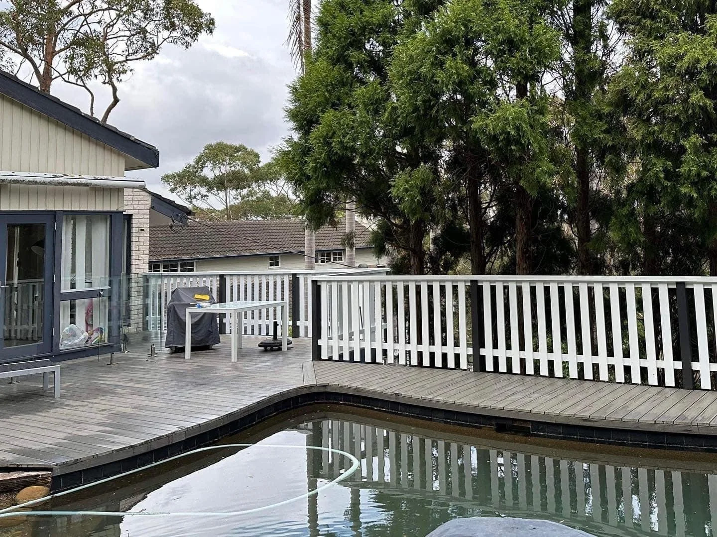 A backyard patio with a wooden deck, a hot tub, a white railing, a black grill, a white table, and trees in the background under overcast sky.