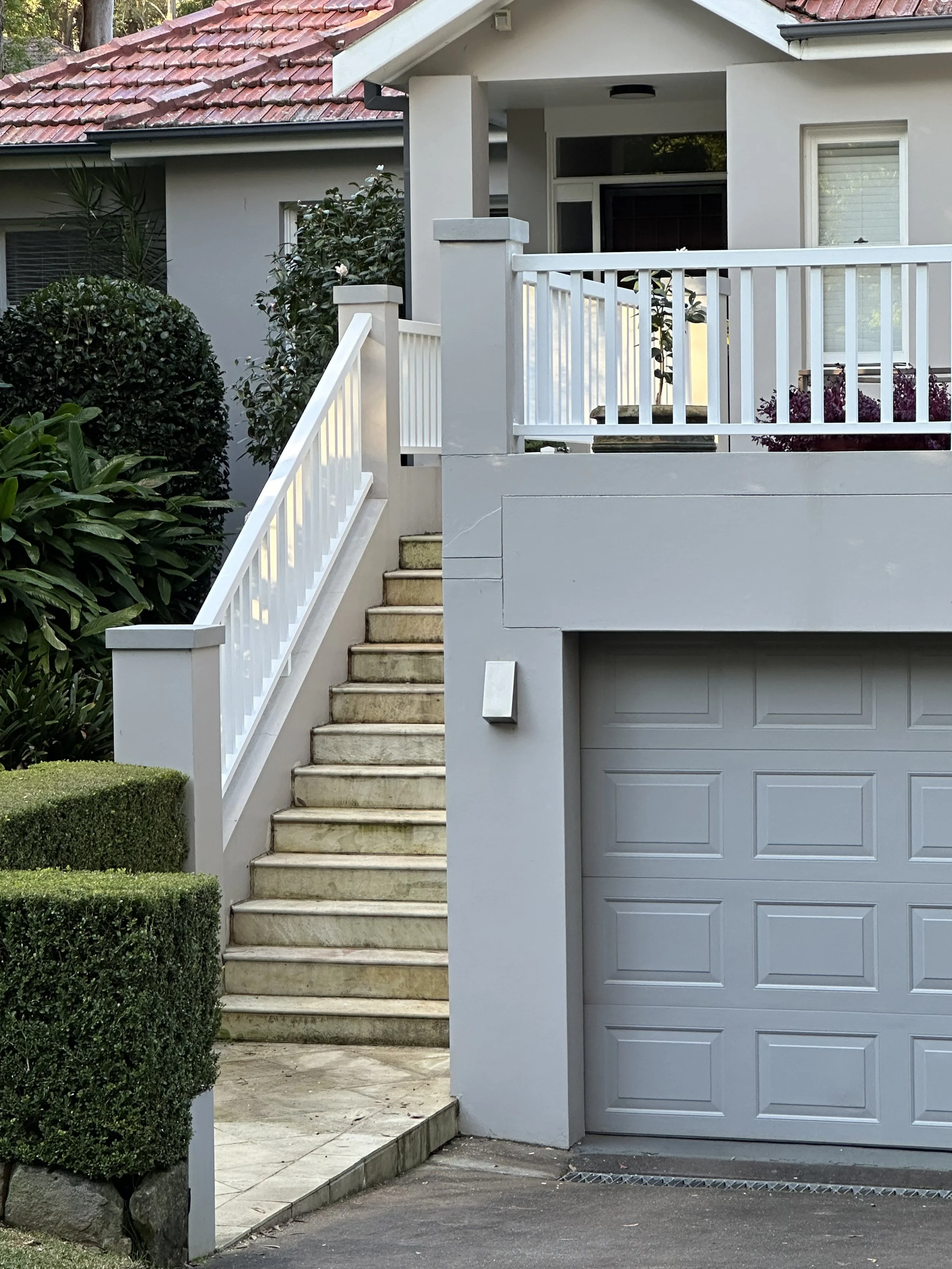 Exterior view of a house showing a stairs with a white railing leading to a porch, a garage door below and lush greenery around.