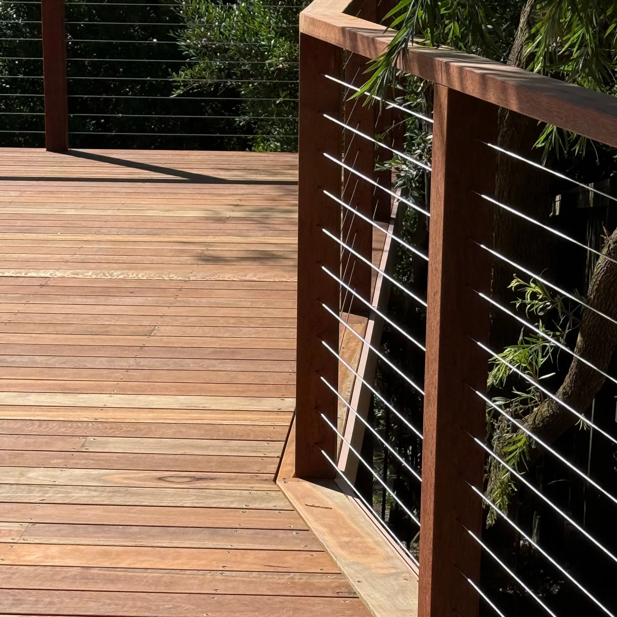 A wooden deck with a railing made of horizontal metal wires and wooden posts, with green foliage in the background, during daytime.
