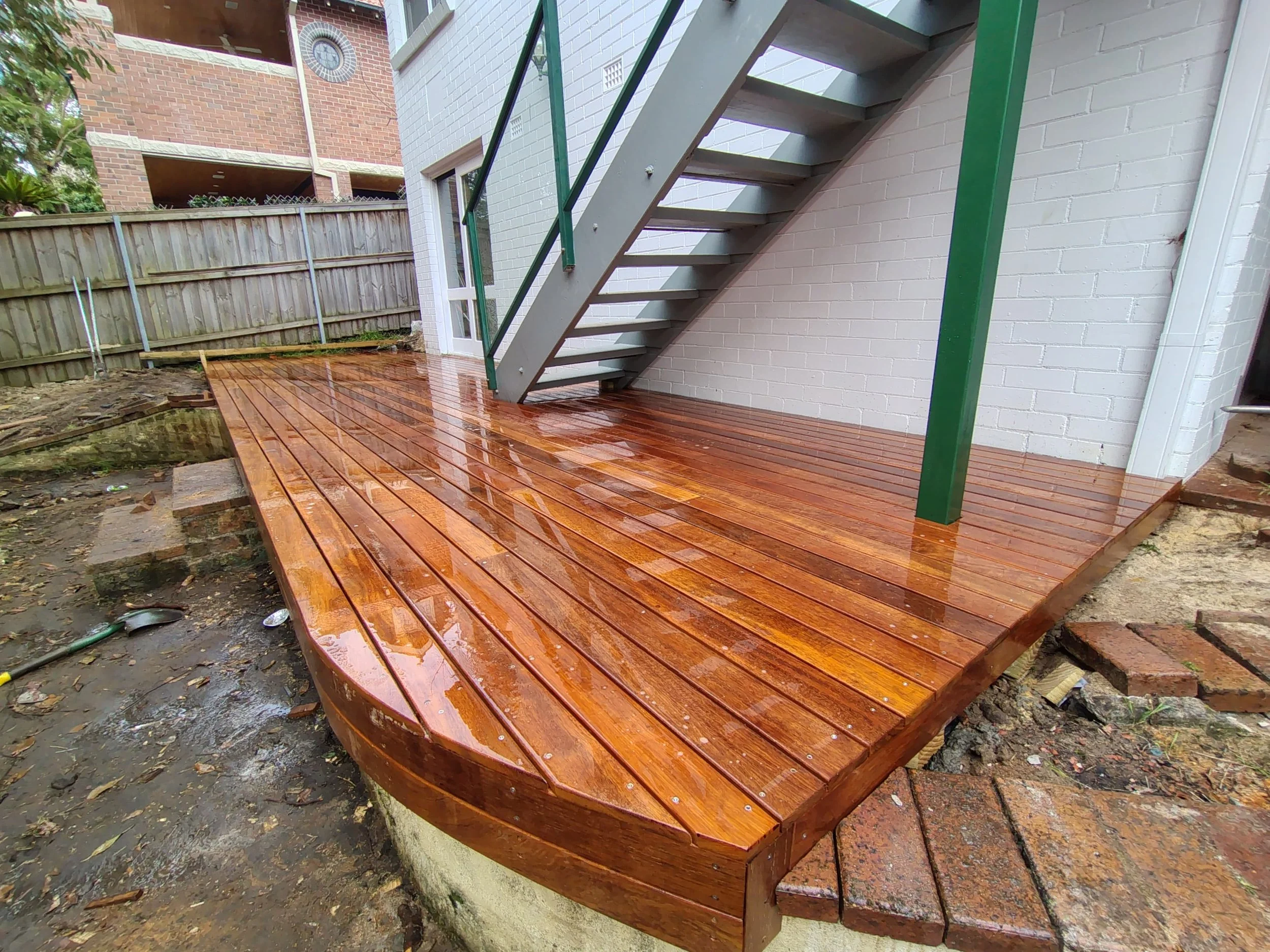 Newly installed wooden deck with rain droplets, attached to a white brick house, next to an exterior staircase with green metal railing.