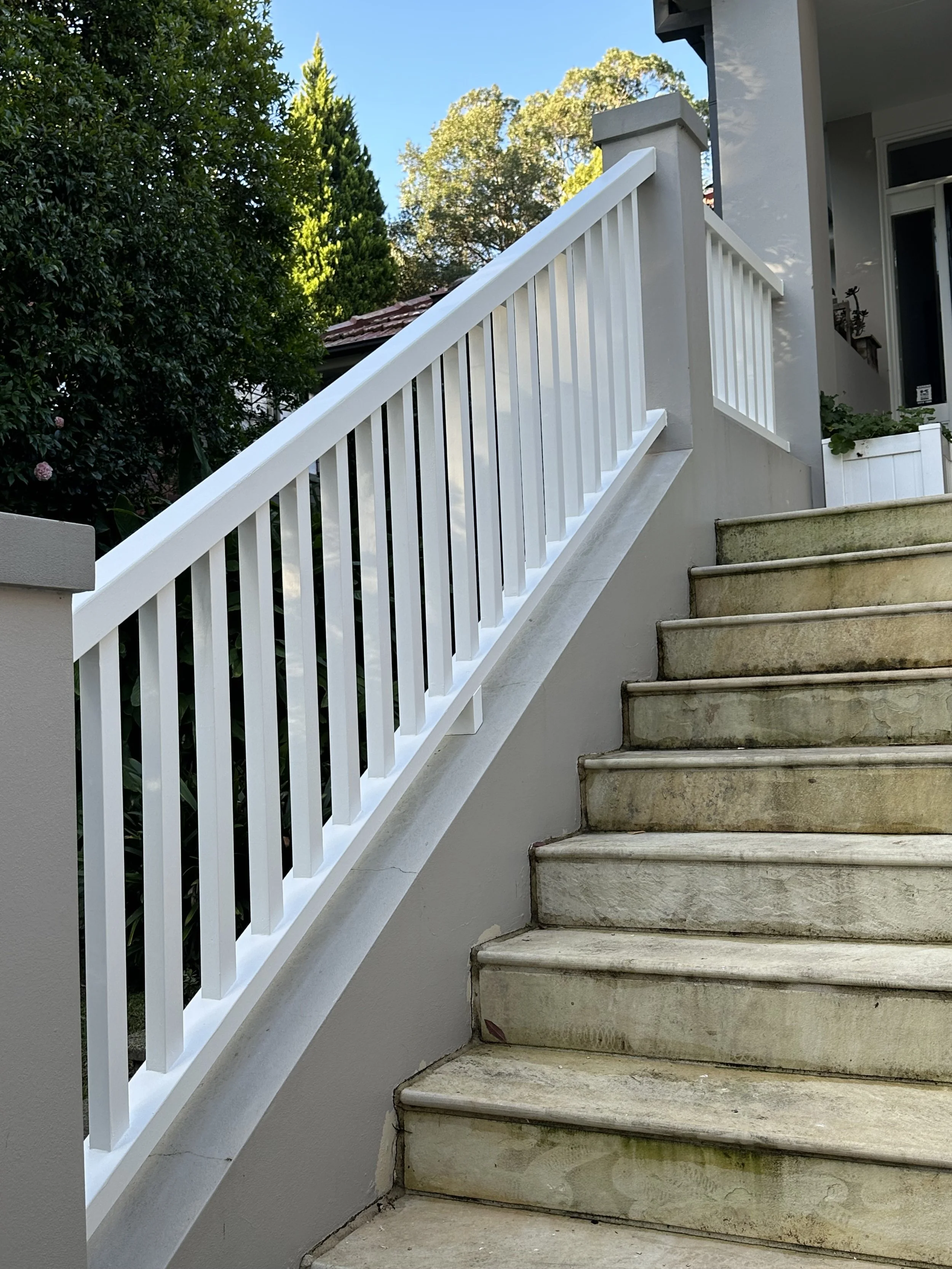 Exterior staircase with beige concrete steps and a white railing, leading up to a house with a porch and greenery in the background.