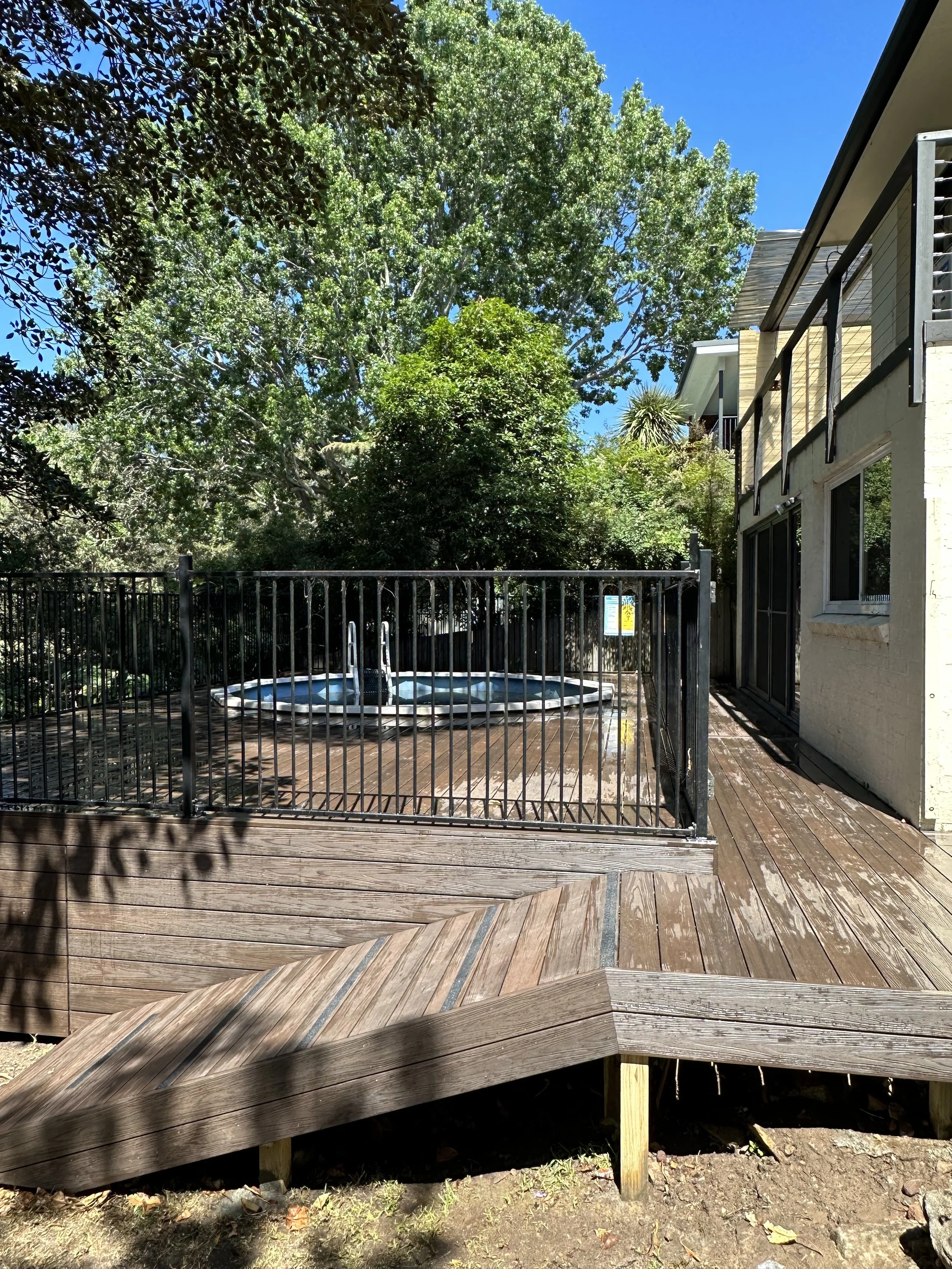 A backyard wooden deck with a hot tub enclosed by a black metal safety fence, surrounded by trees and a house with a balcony on a sunny day.