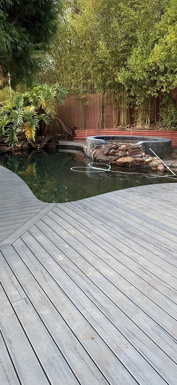 A backyard with a wooden deck, a small pond with dark water, and a hot tub surrounded by rocks and a wooden fence, with trees and bushes in the background.