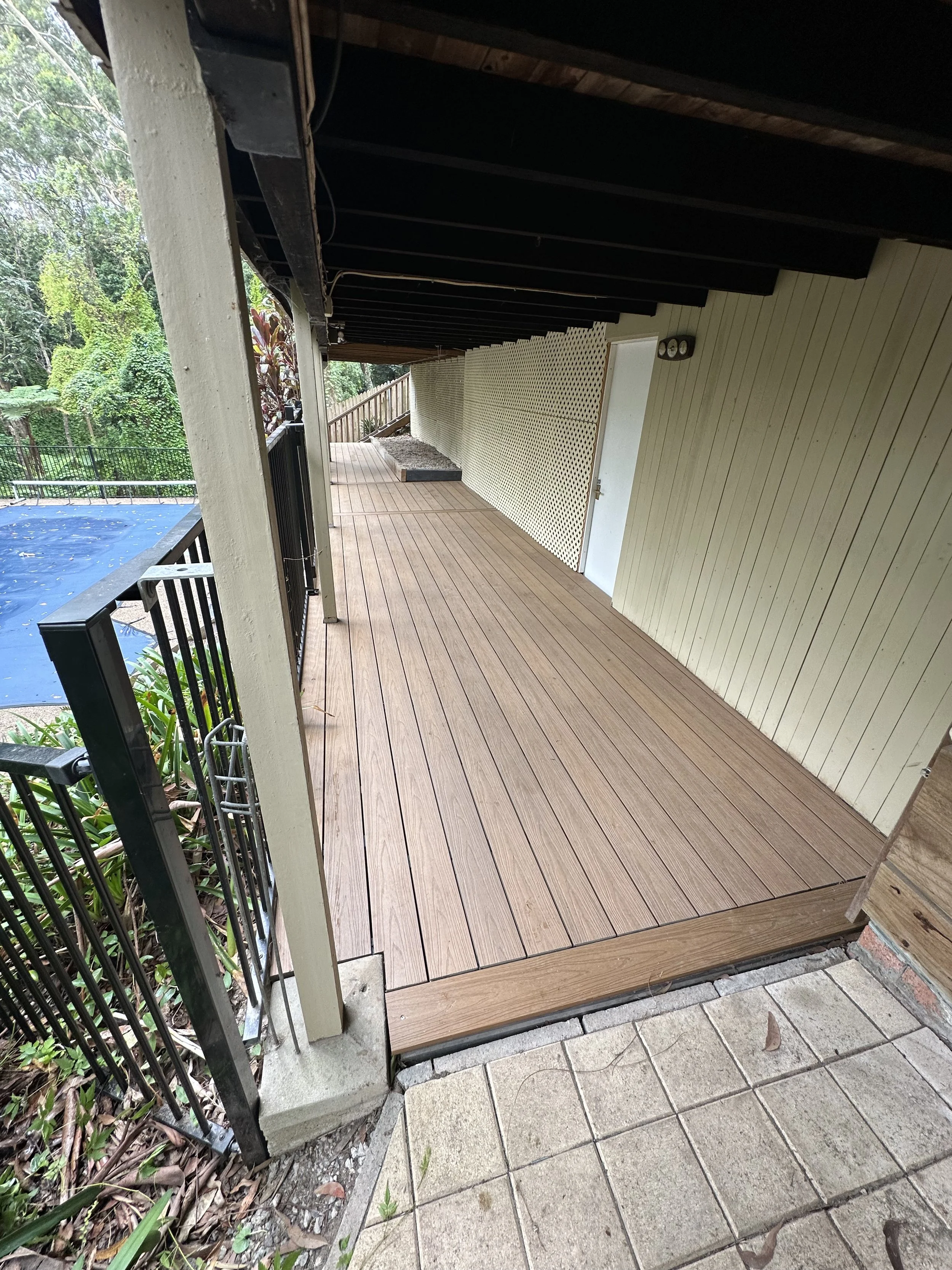 Wooden deck area with black metal railing, a door on the wall, and surrounding greenery.