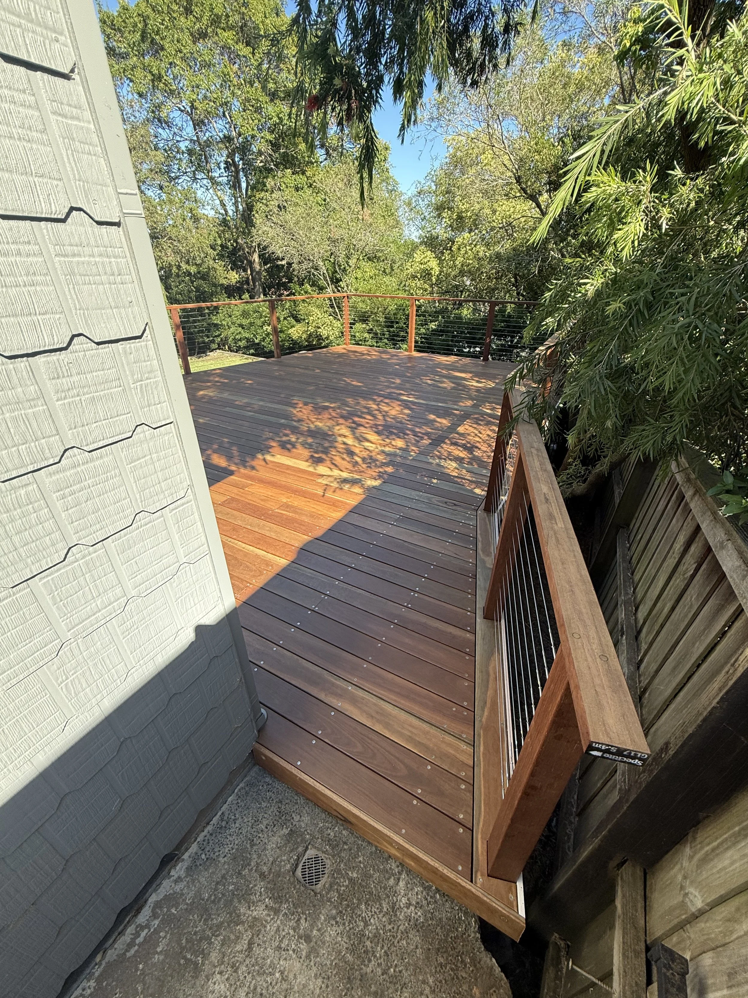 A freshly built wooden outdoor deck with a railing, surrounded by trees and outdoor greenery, with sunlight casting shadows on the surface.