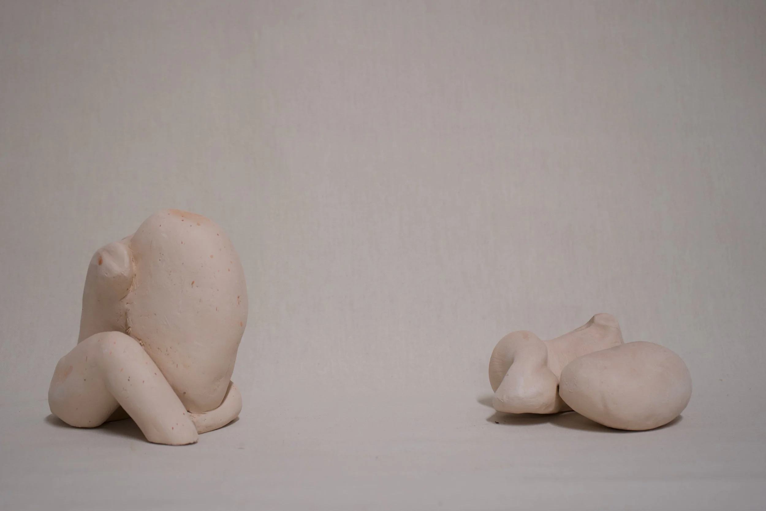 Zwei Skulpturen aus weißem Ton, eine auf der linken Seite mit einer abstrakten menschlichen Form, die auf Knien sitzt, und die andere auf der rechten Seite mit einer kleineren, unauffälligen Form, auf einem neutralen Hintergrund.
