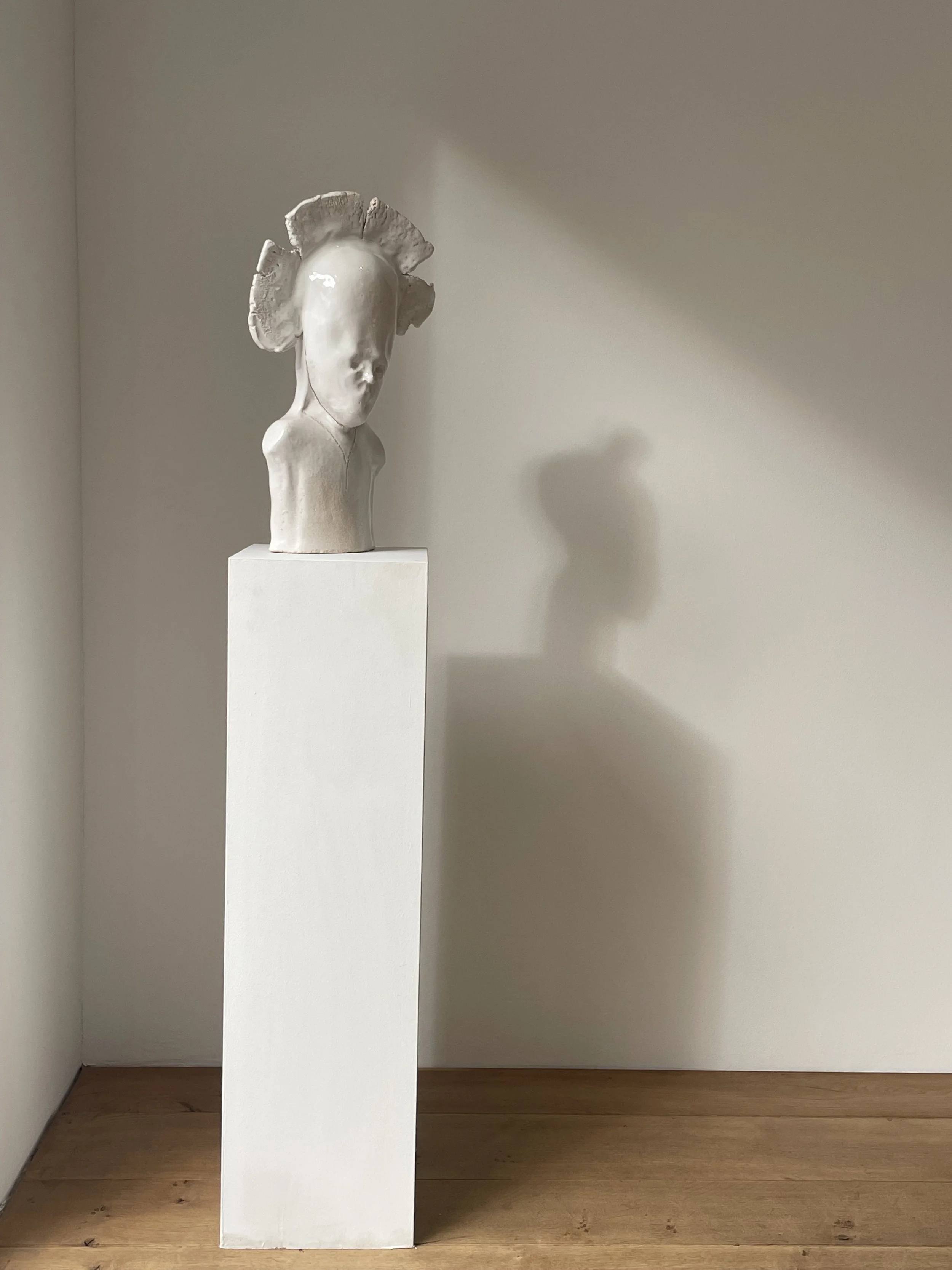 Skulptur eines menschlichen Kopfes mit einer modifizierten Frisur auf einem weißen Sockel, auf einem Holzboden, Schatten an der Wand.
