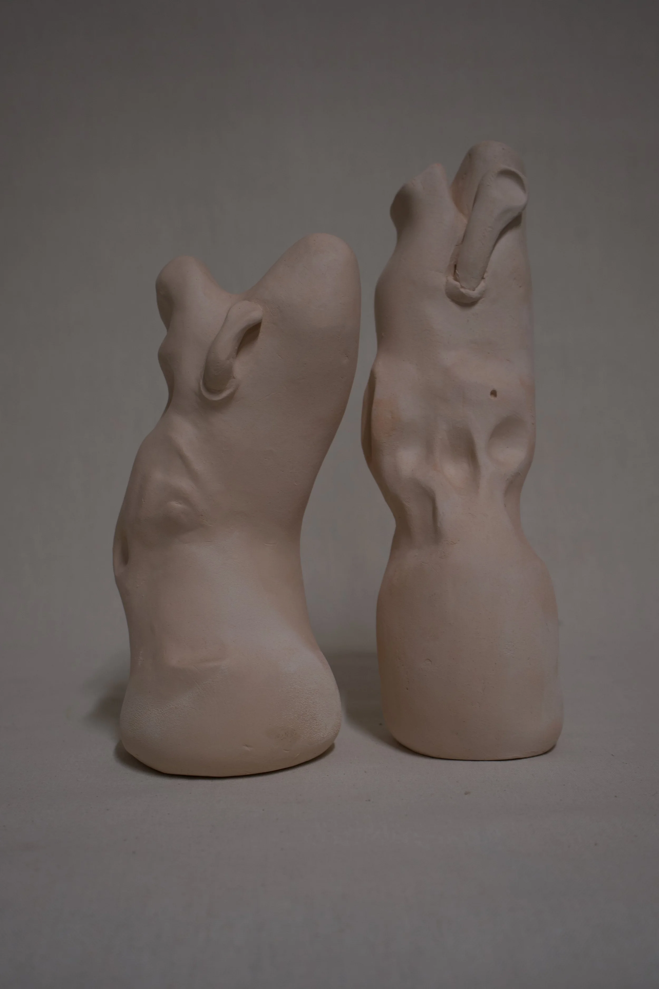 Zwei skulpturale Köpfe aus Ton, die menschliche Gesichter auf abstrakte Weise darstellen, mit hervorstehenden Ohren und offenen Mündern.