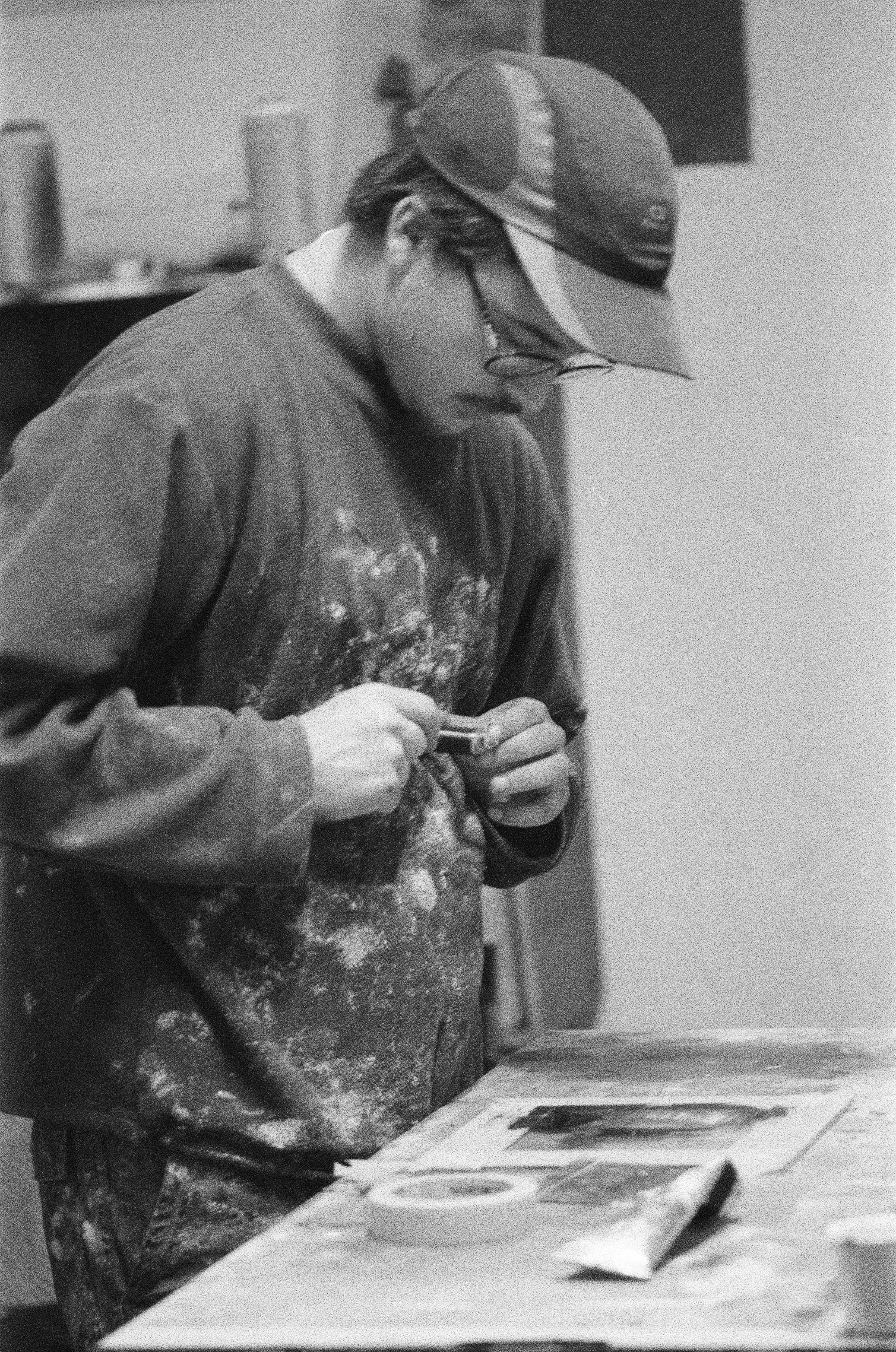 Junge Person trägt eine Mütze und Brille, beschäftigt sich mit einem Smartphone, trägt eine verschmutzte, kaputte dunkle Jacke, sitzt an einem Tisch, auf dem sich Papiere, eine Schere und ein Päckchen befinden.