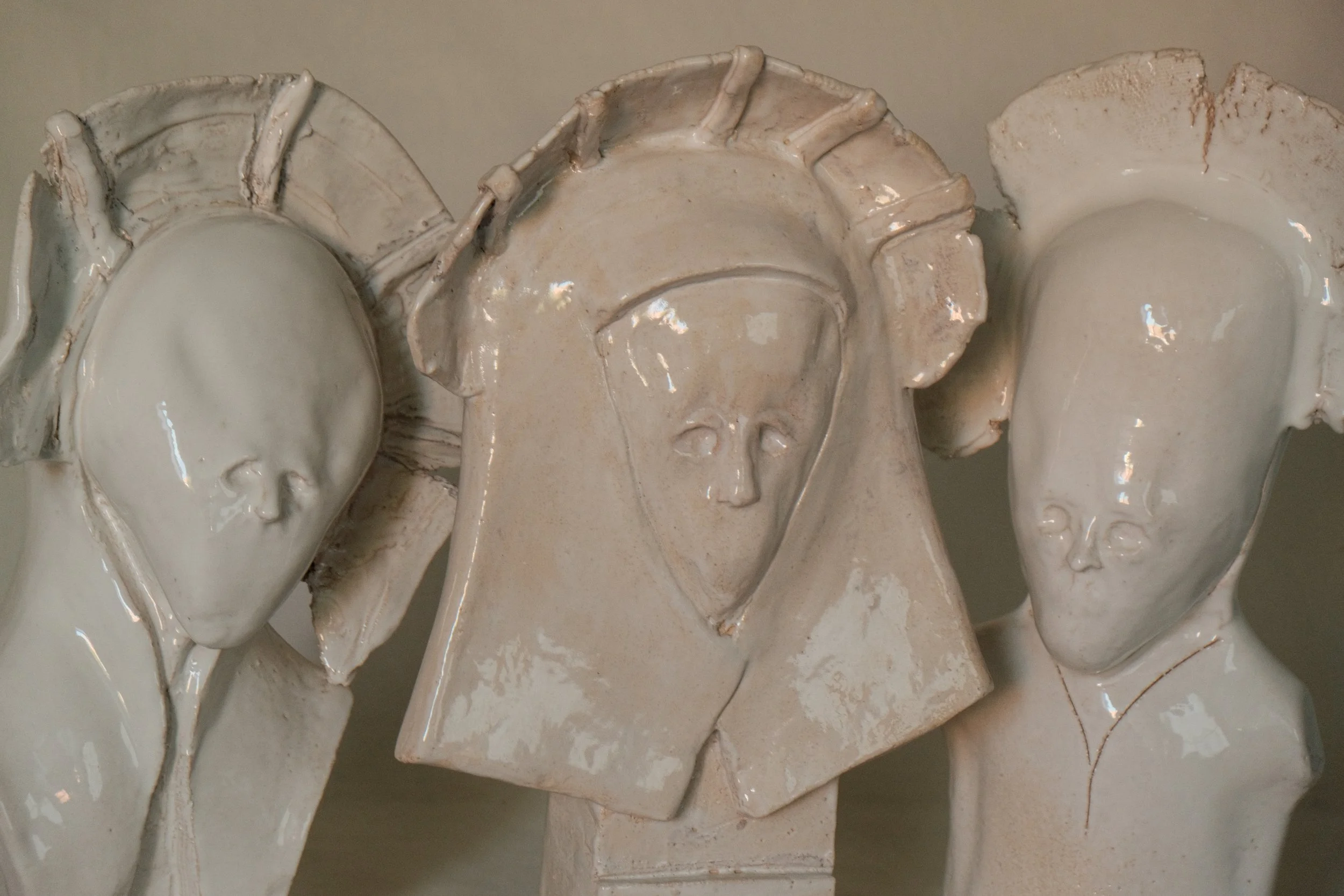 Drei weiße Keramikskulpturen mit menschlichen Gesichtern und Konturen, die an Heilige oder Figuren mit religiösem Hintergrund erinnern.