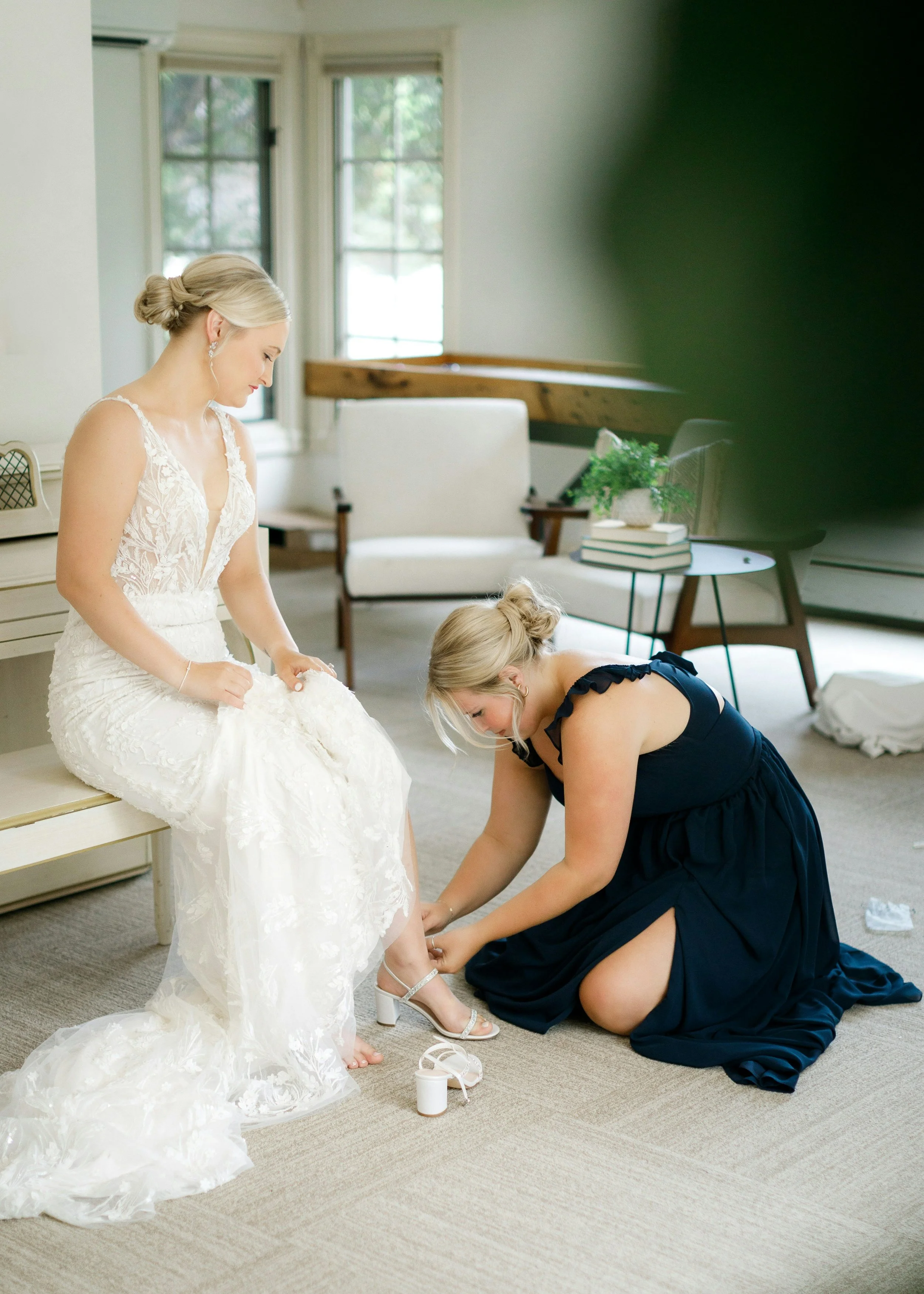 Bride getting ready on wedding morning while a bridesmaid helps fasten her wedding shoes.