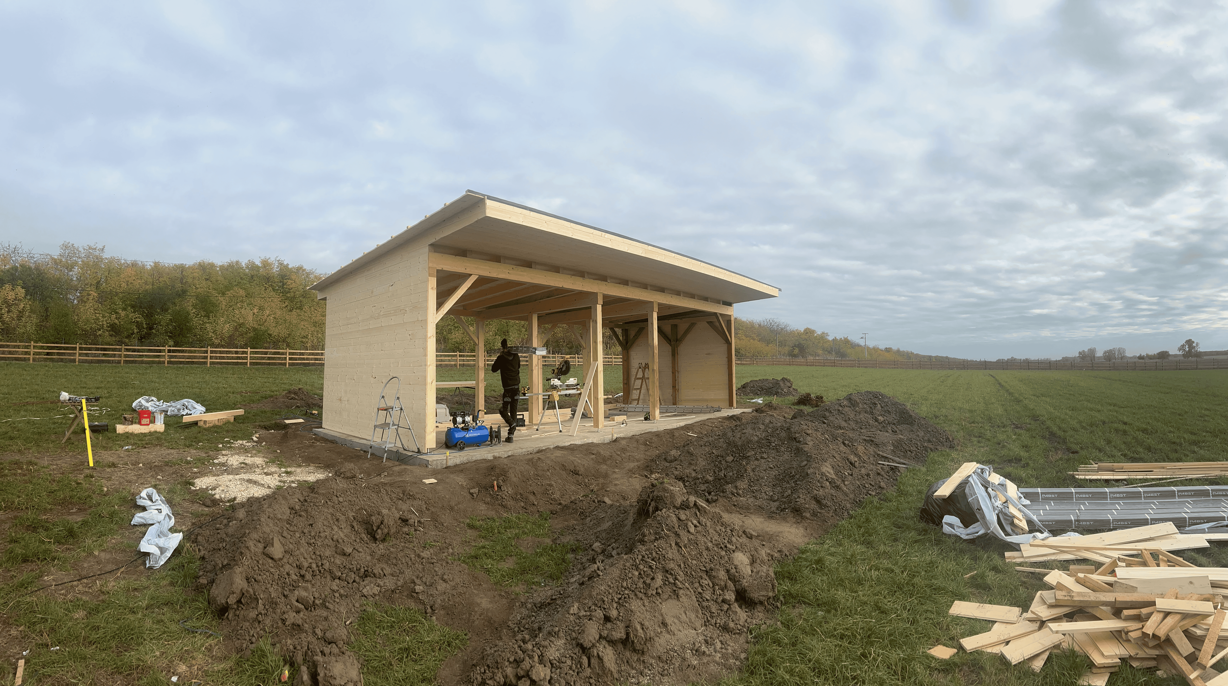 Construction of a small wooden shed in a grassy field with tools and materials scattered around on a cloudy day.
