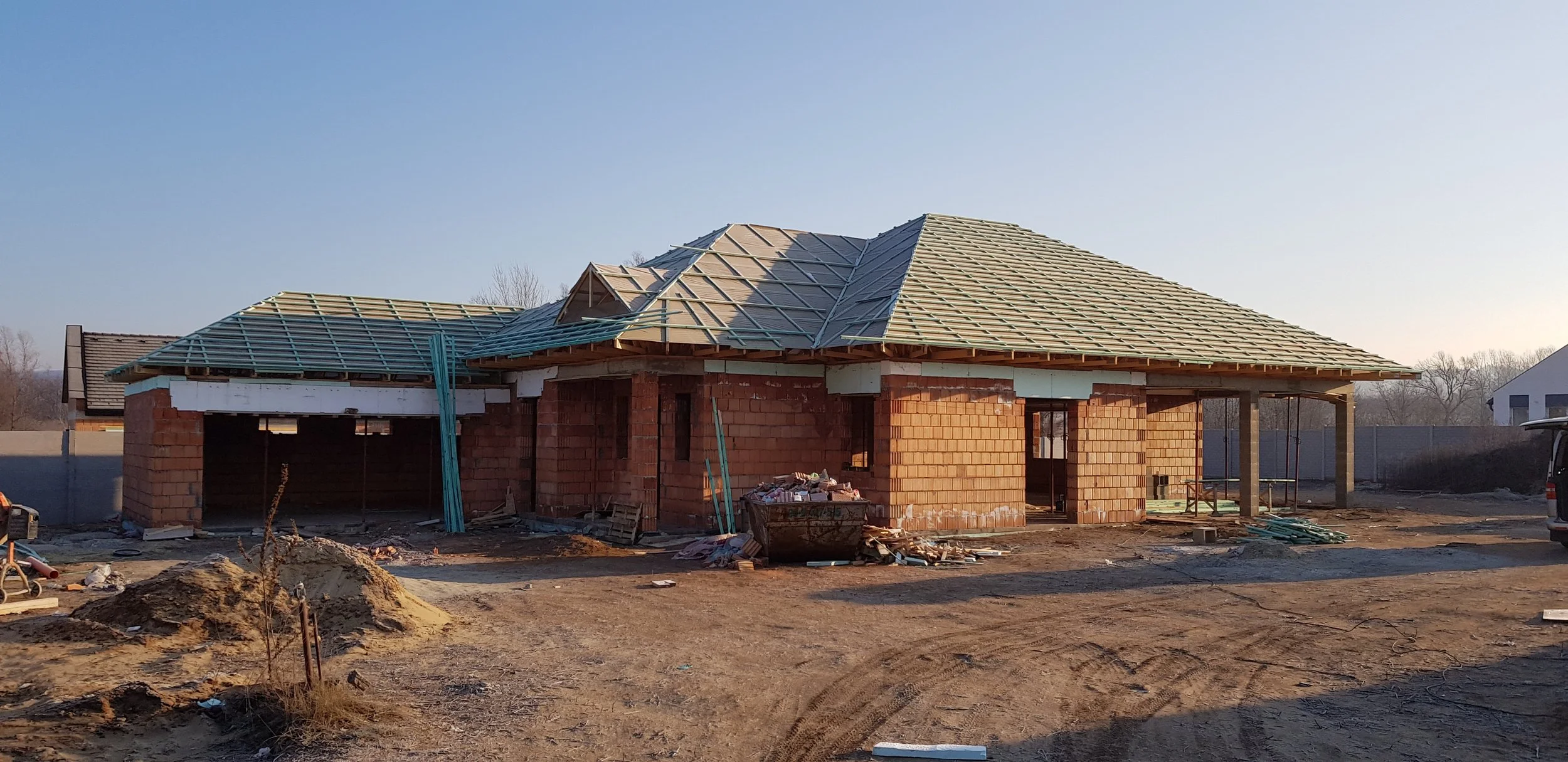 A house under construction with a brick exterior and a partially completed roof, surrounded by construction materials and dirt ground.