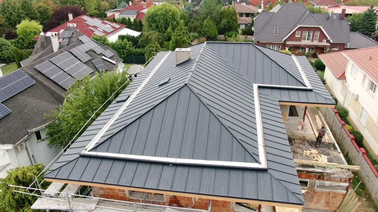 A bird's eye view of a house with a dark gray metal roof, under construction, with construction materials and a worker on site. Surrounding houses have various roof styles, including solar panels and different colors.