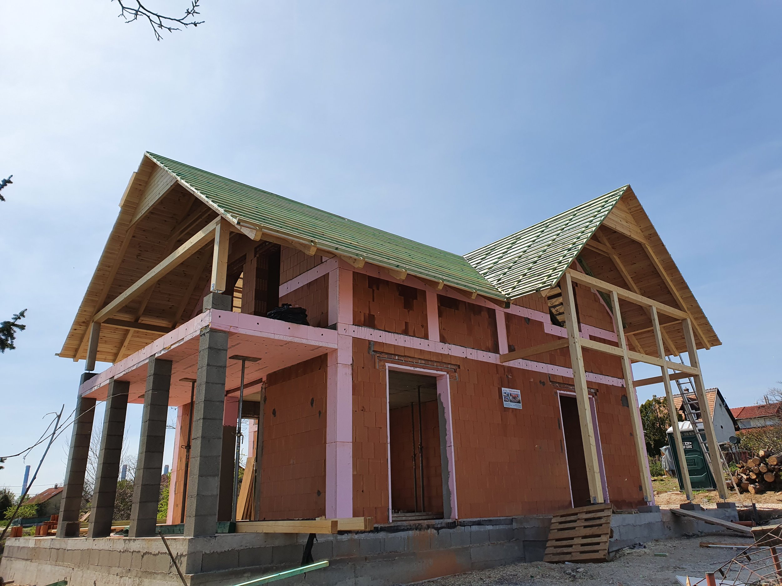 A two-story house under construction with brick walls, wooden framing, and green roof shingles, on a concrete foundation, in a residential area with trees and other houses.