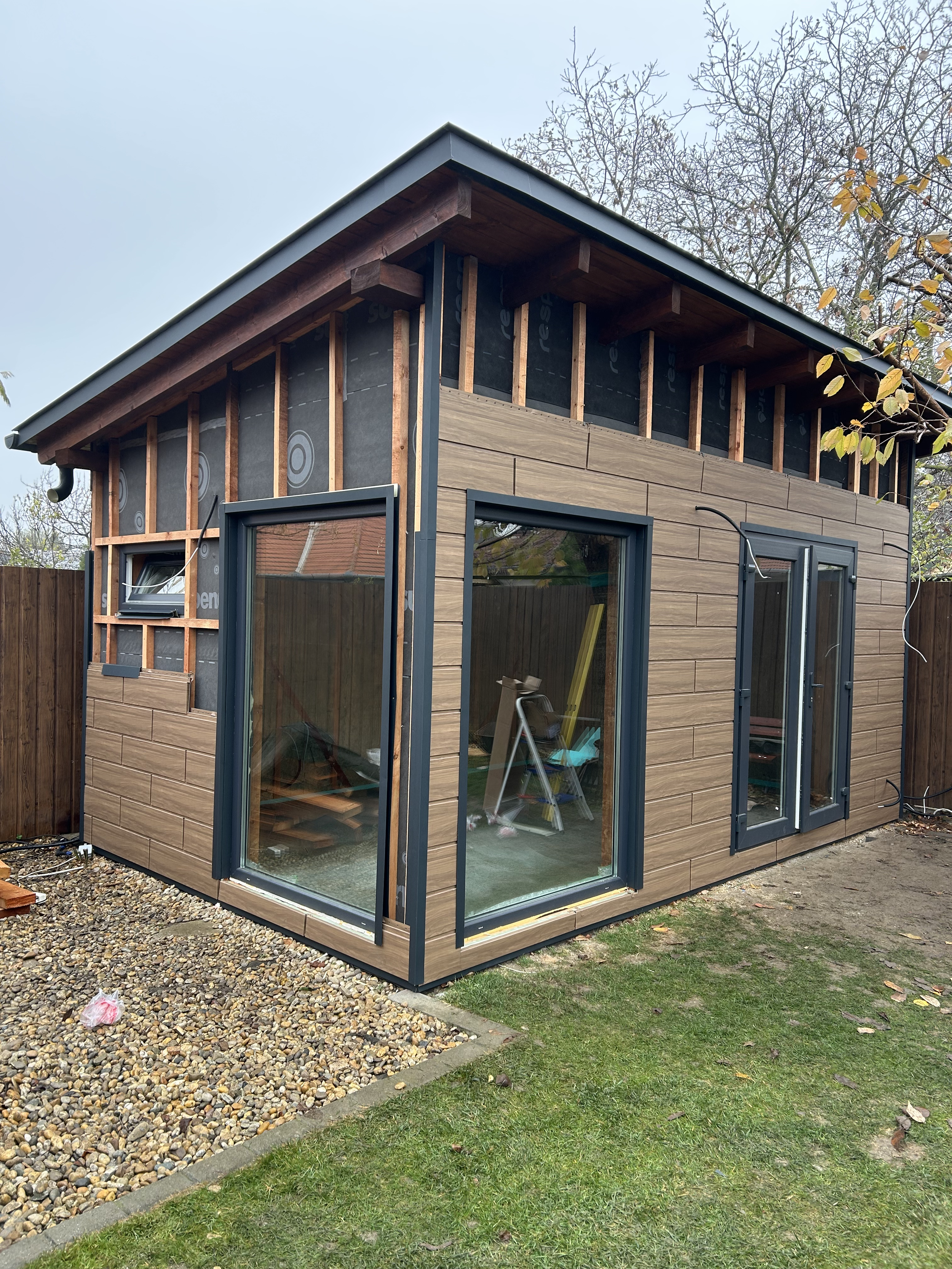A small modern house under construction with wood siding and large glass sliding doors, surrounded by a grassy yard and fenced backyard.
