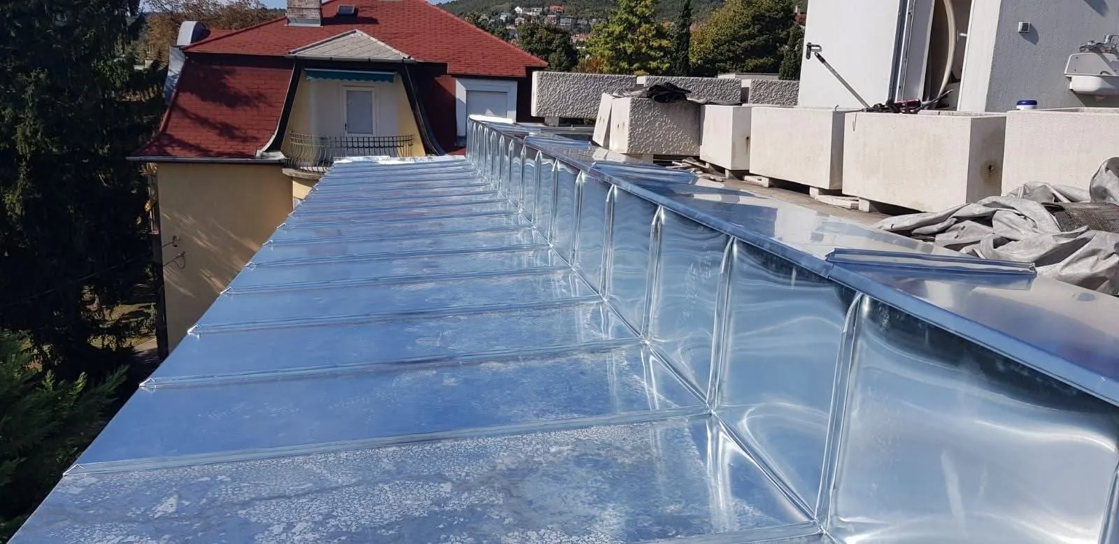 Construction site on a roof with shiny aluminum or metal panels, with houses and trees in the background.