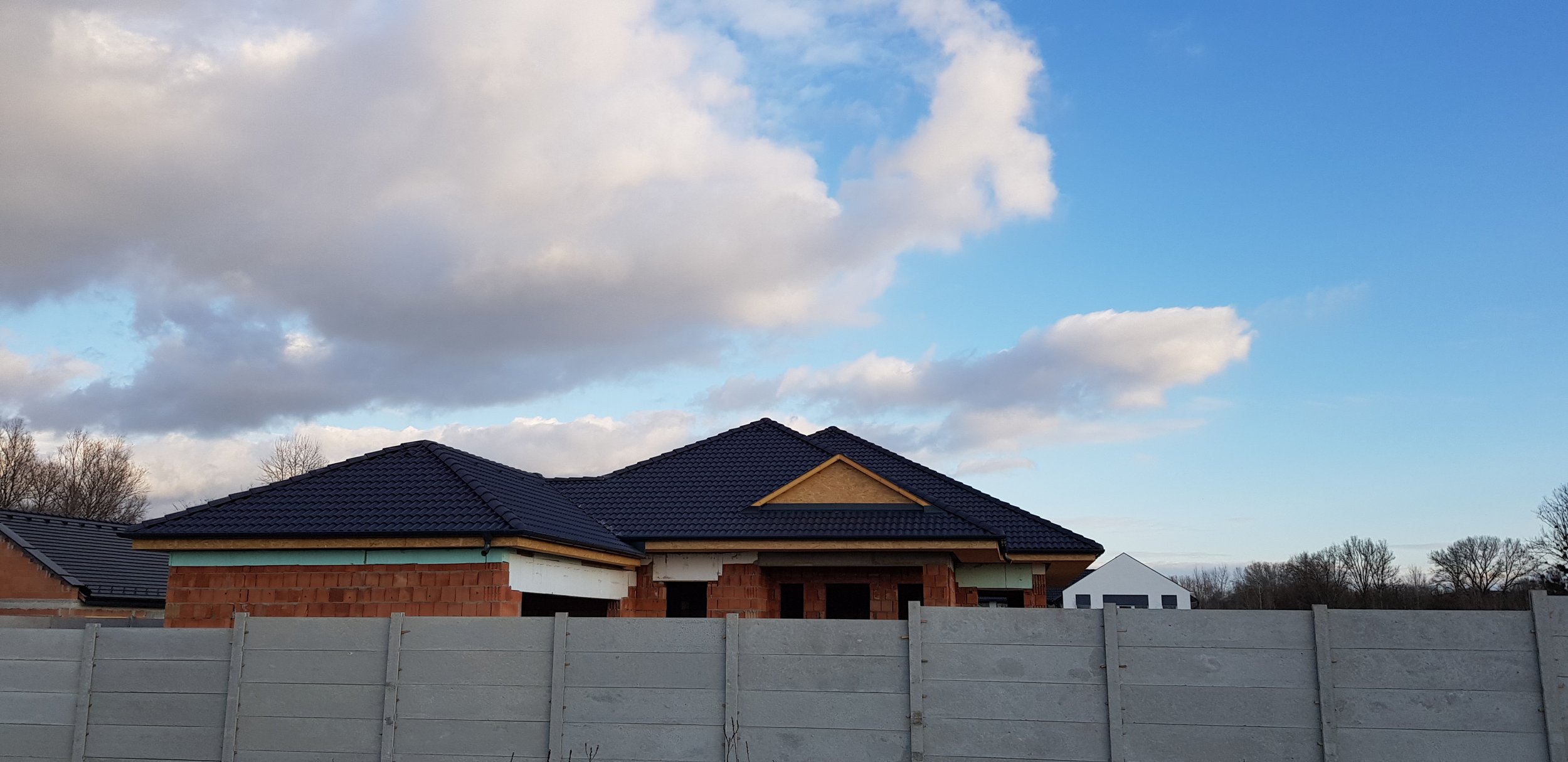 Under construction house with partially built brick walls and new dark gray tiled roof, surrounded by a gray fence, under a partly cloudy sky.