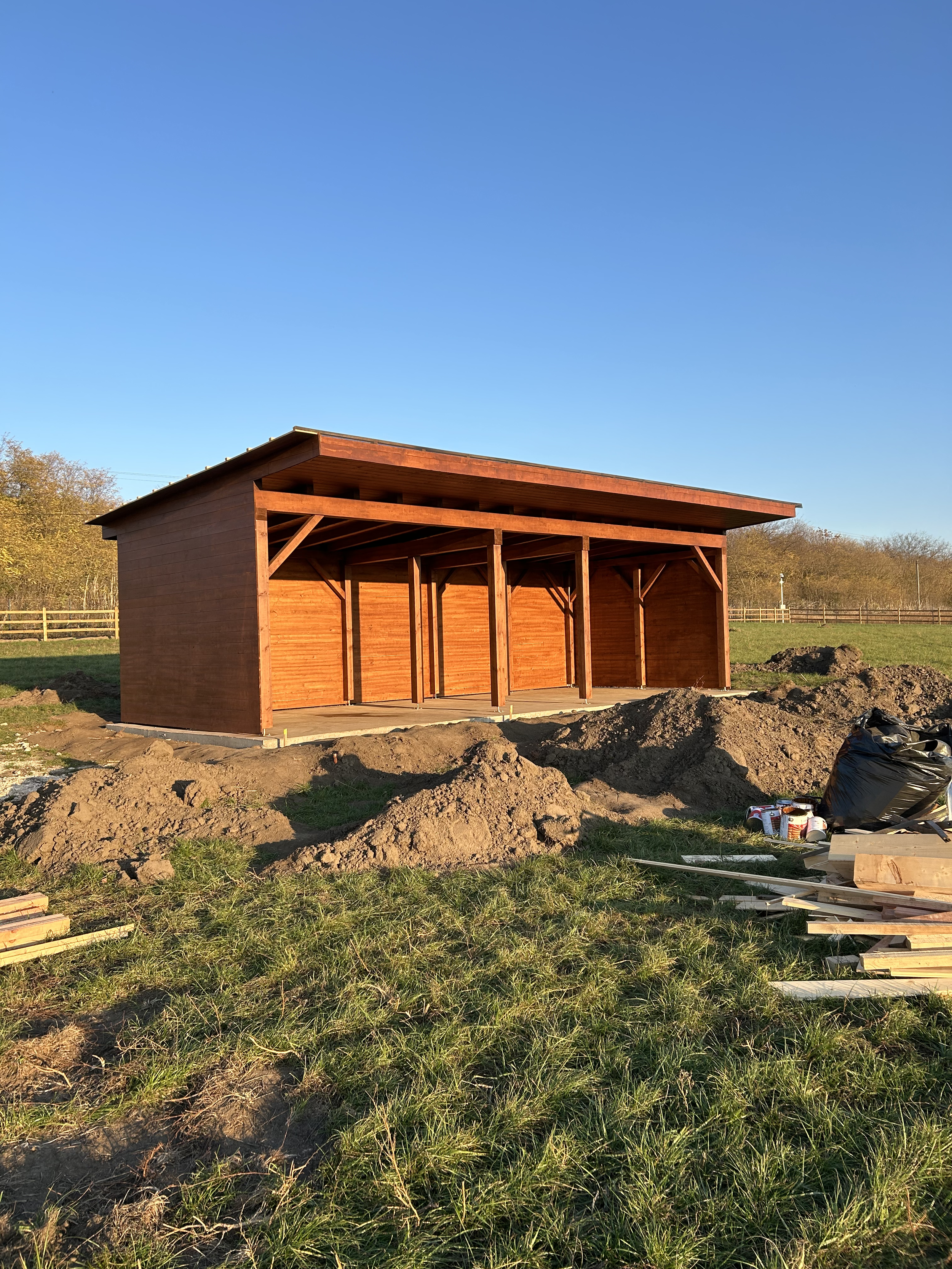 A wooden shed under construction on a grassy field with piles of dirt, construction materials, and tools nearby, under a clear blue sky.
