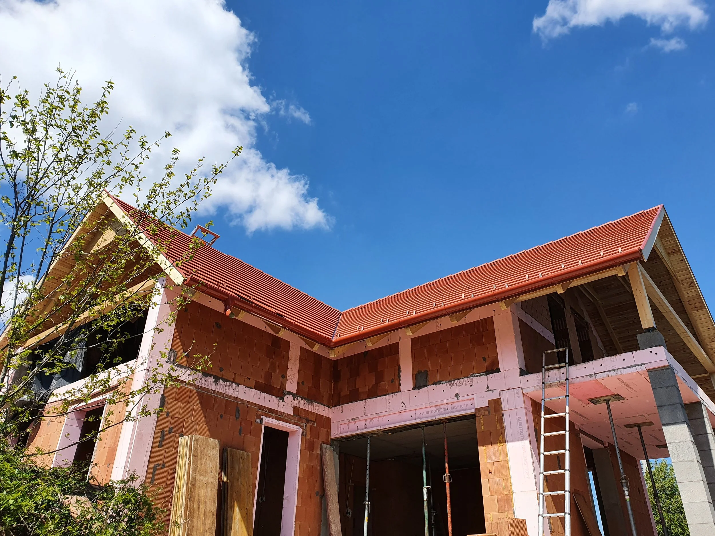 A two-story house under construction with a red-tiled roof, exposed red brick walls, a ladder leaning against the house, and a partly cloudy blue sky in the background.