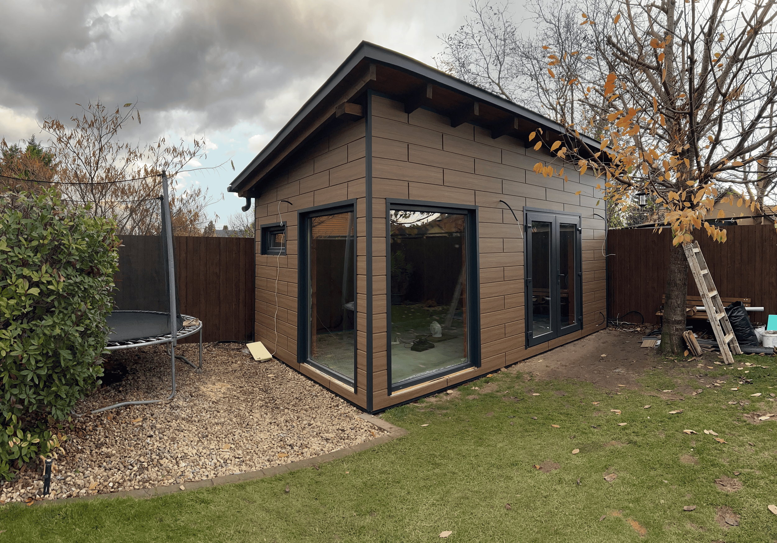 Small modern backyard shed with large glass doors and wood siding, surrounded by a lawn, trees, and fencing, with some construction tools and materials nearby.