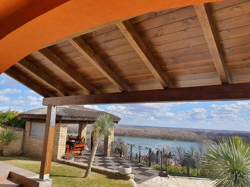 View from a covered patio overlooking a river with trees on the opposite bank, featuring a small brick structure with a thatched roof and outdoor furniture, landscaped with plants and a decorative fence.