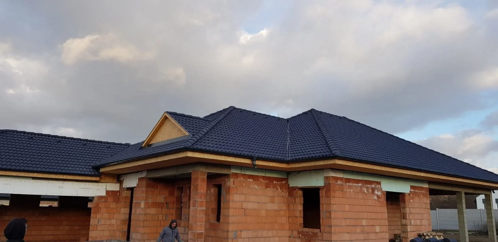 House under construction with a newly installed dark blue tiled roof, brick walls, and construction workers present.