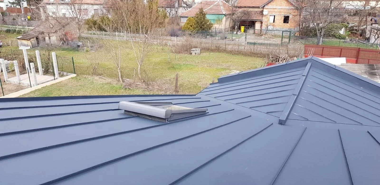 View of a metallic gray roof with a skylight window, overlooking a backyard with trees, grass, and neighboring houses.