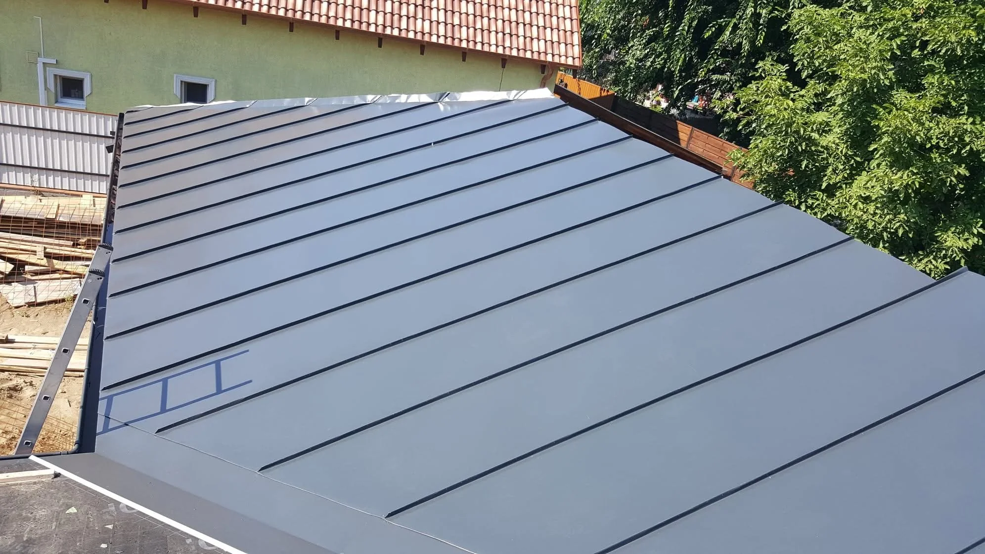 Metal roof panels installed on a sloped roof in a residential area. There is a ladder on the left side, and surrounding trees and houses are visible.