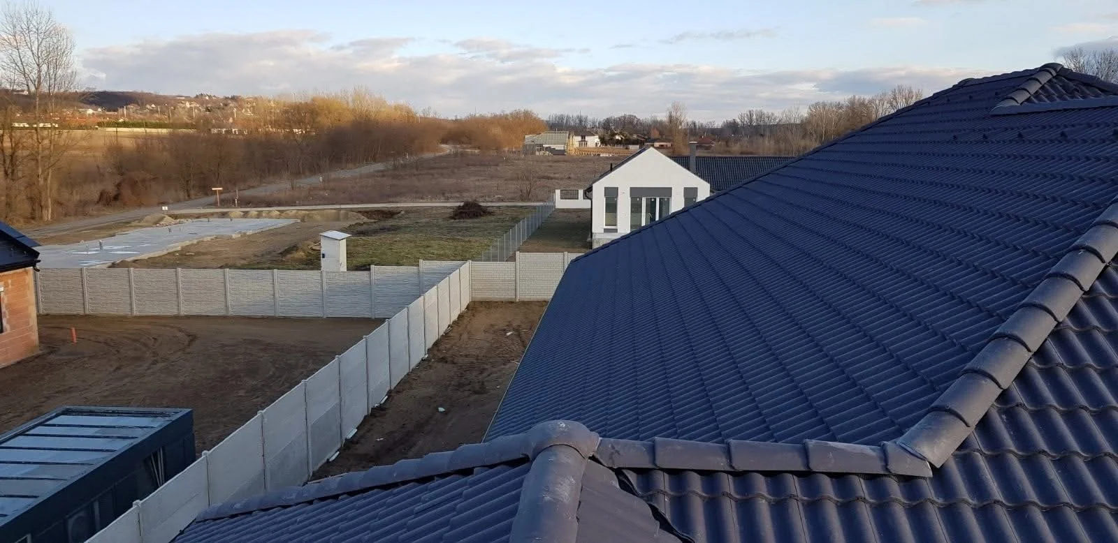 View of rooftops with blue tiled roofing, fenced backyard under construction, and distant landscape with trees and buildings.
