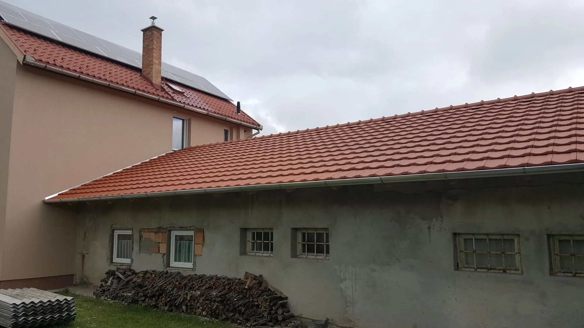 A house with new red roof tiles, an unfinished side with small barred windows, and an adjacent taller building with solar panels and a chimney.