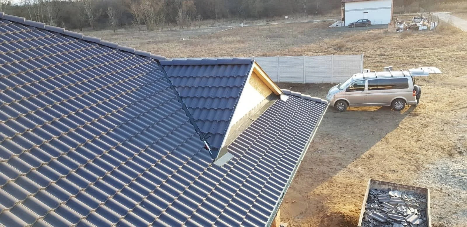 View of a house roof with black solar panel tiles, a pickup truck with solar panels on top, and a yard with construction materials and equipment.