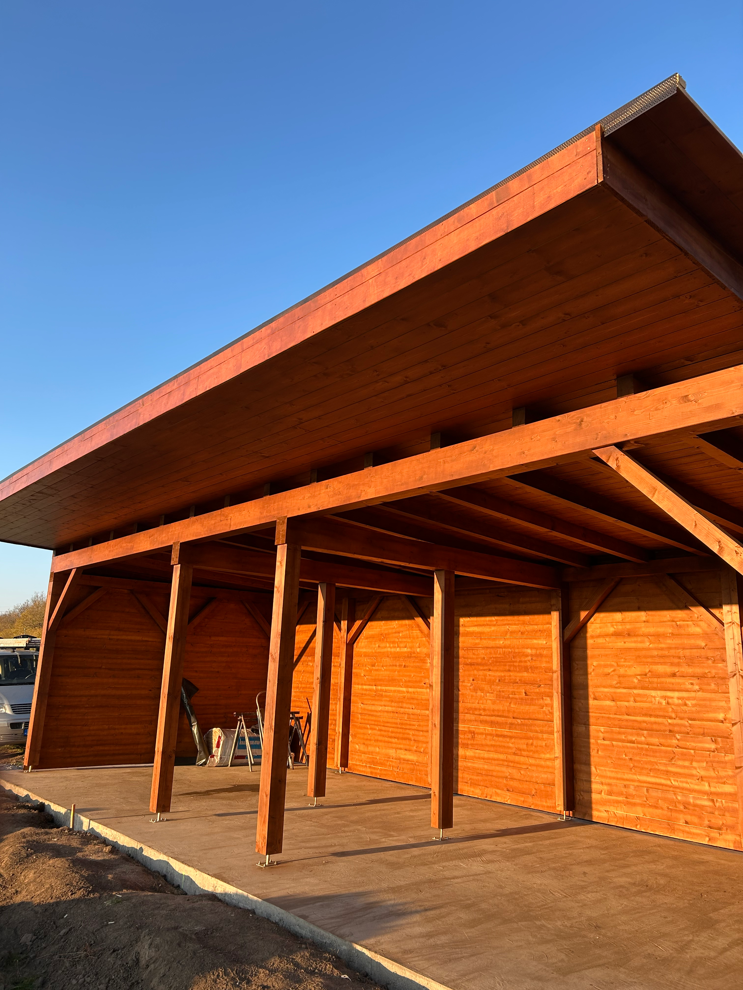 A wooden outdoor structure under construction with supporting beams, a concrete foundation, and a roof made of wooden planks, set against a clear blue sky.