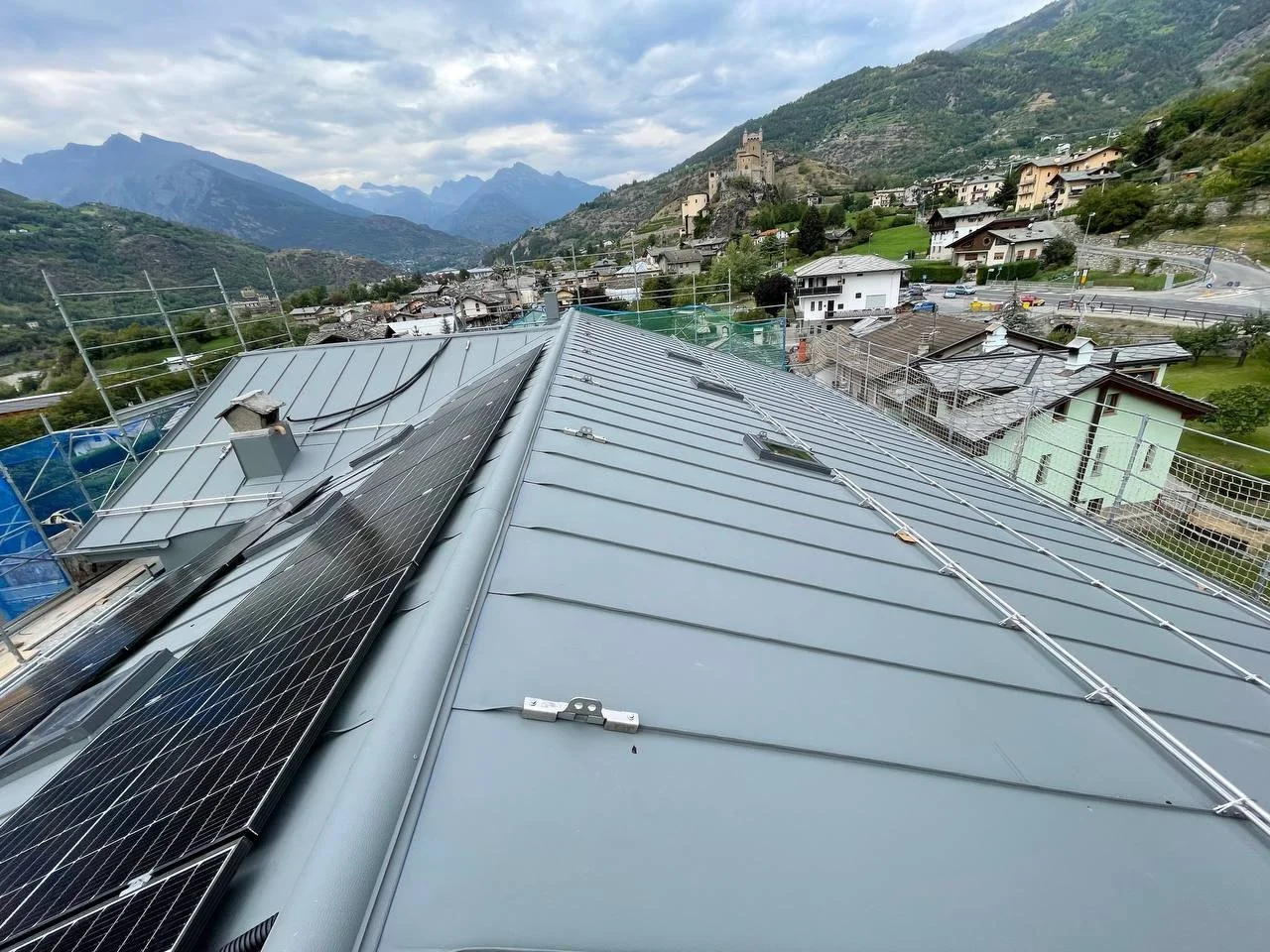 Vista di un tetto in metallo con pannelli solari e moduli fotovoltaici, sopra un paesaggio di montagna con case e un castello in lontananza.